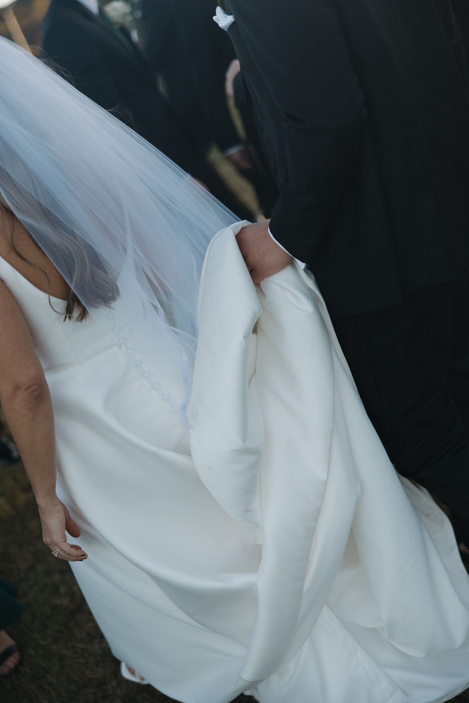 Close-up of a bride in a white satin wedding gown with veil, holding her dress train at an outdoor wedding.