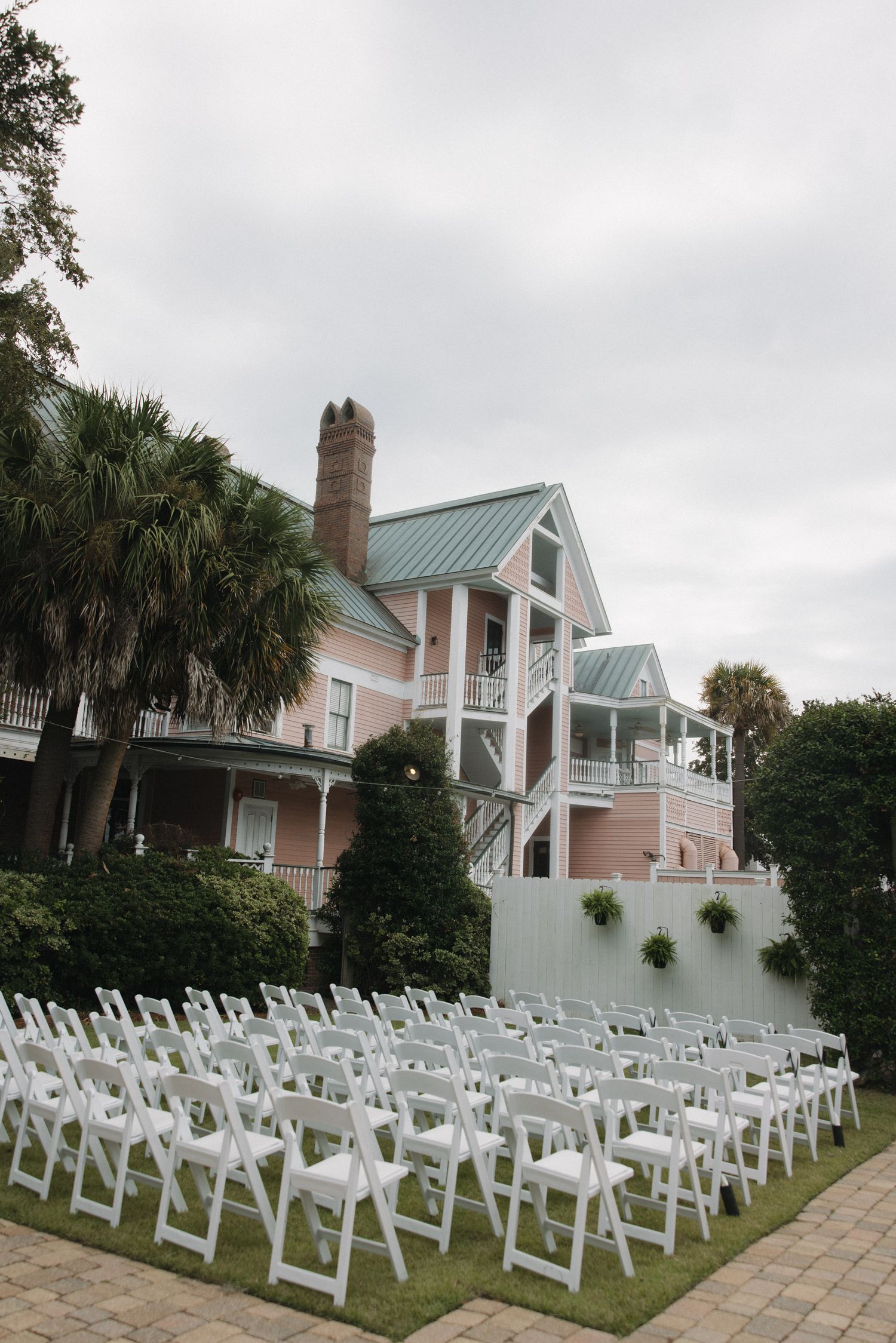 White chairs arranged on a lawn for an outdoor wedding ceremony in front of a pink Southern mansion.