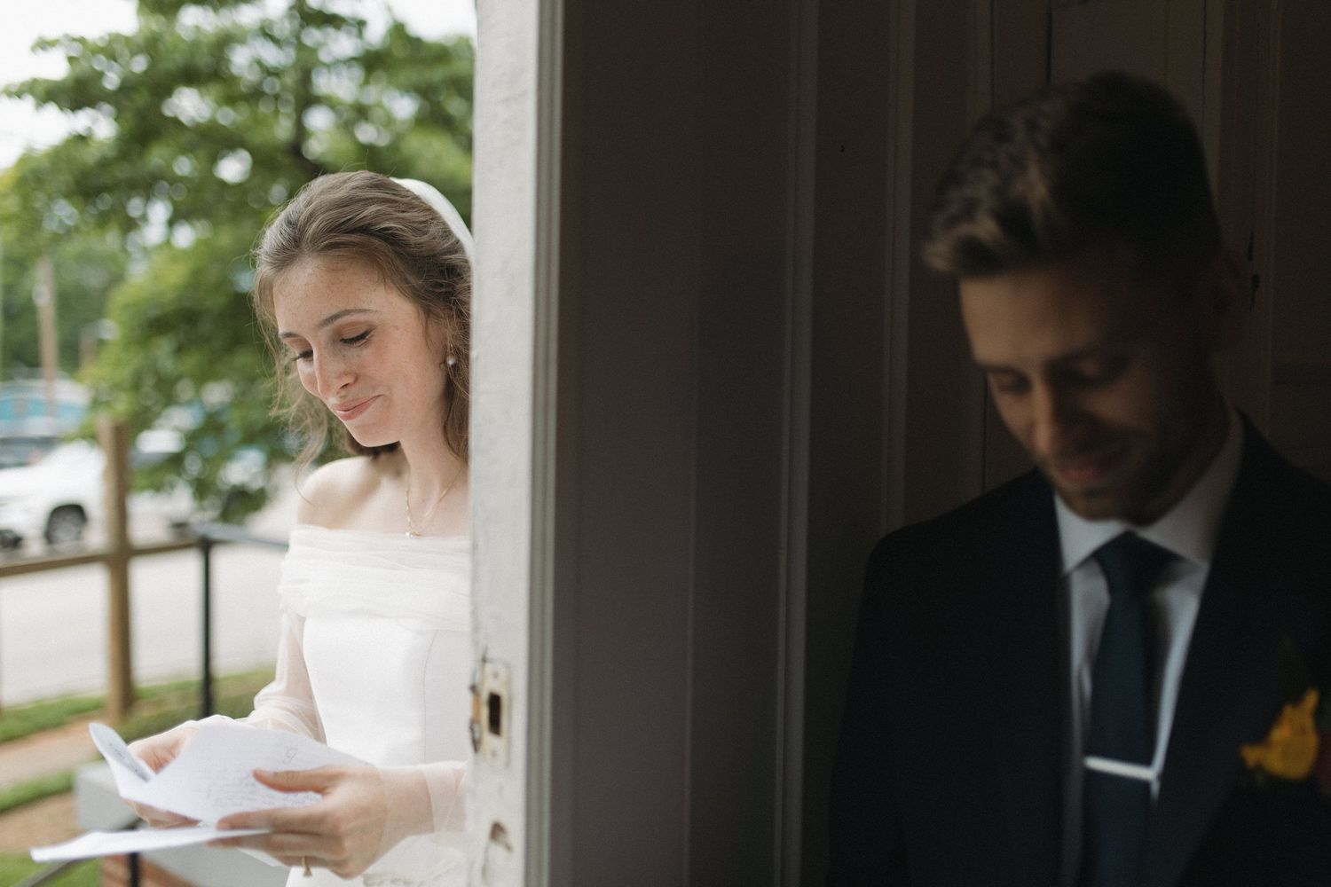 Bride reads a letter beside a door while blurred groom stands on the other side.