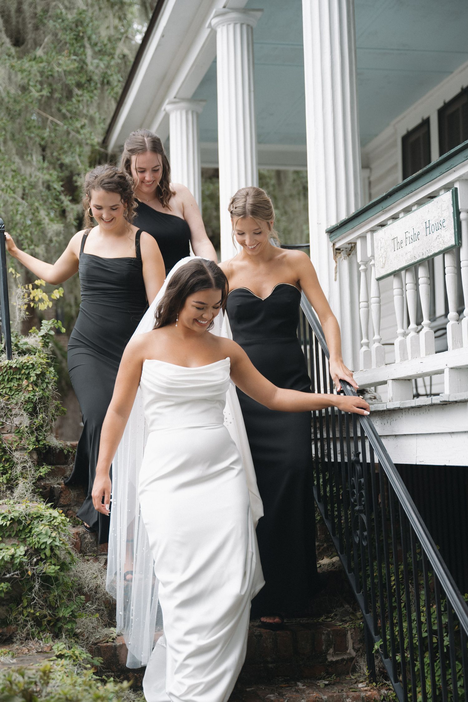 Bride in white strapless gown descending porch steps with bridesmaids in black dresses at historic white house venue.