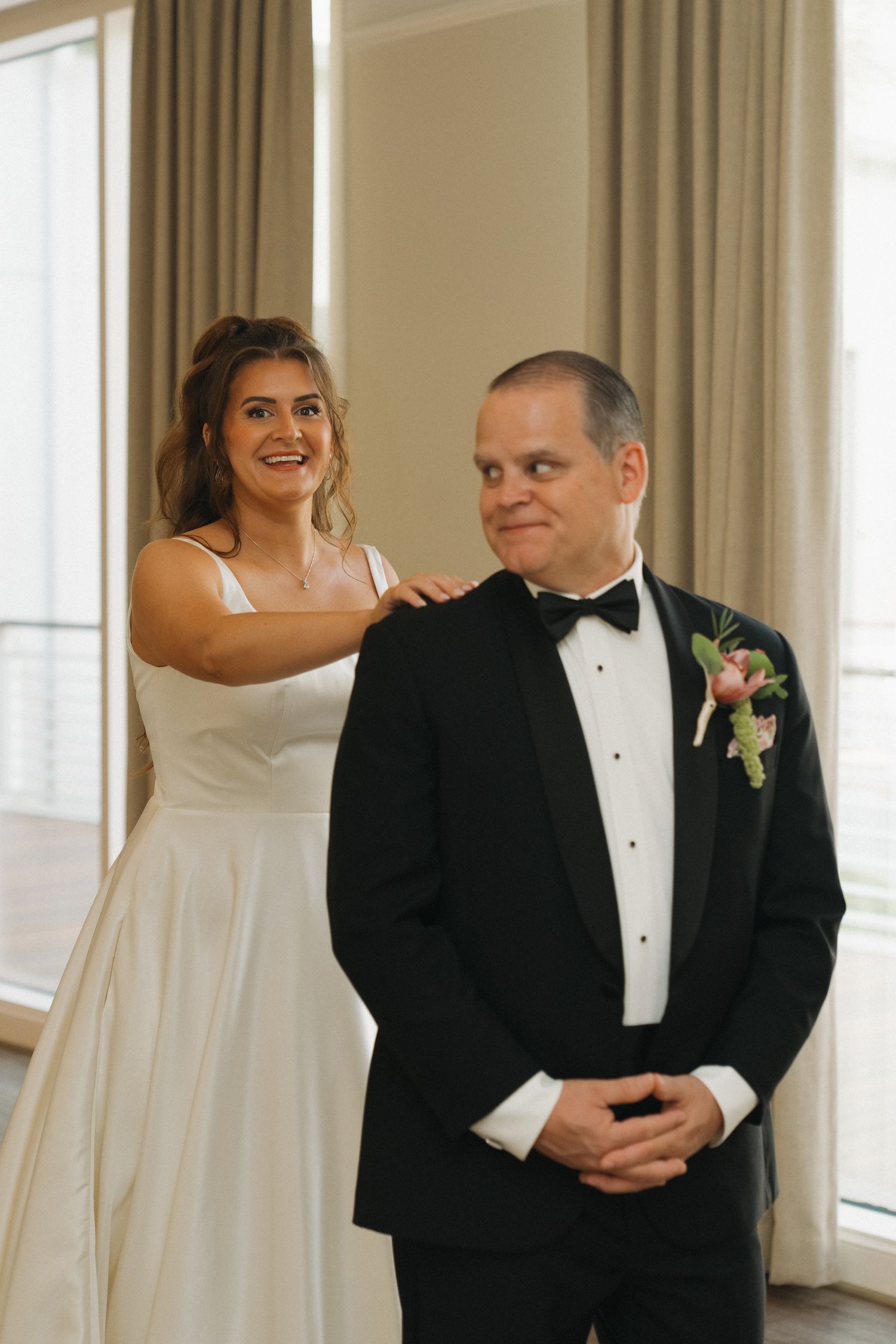 Bride in white gown taps groom's shoulder during first look moment, groom in black tuxedo with floral boutonniere smiles.