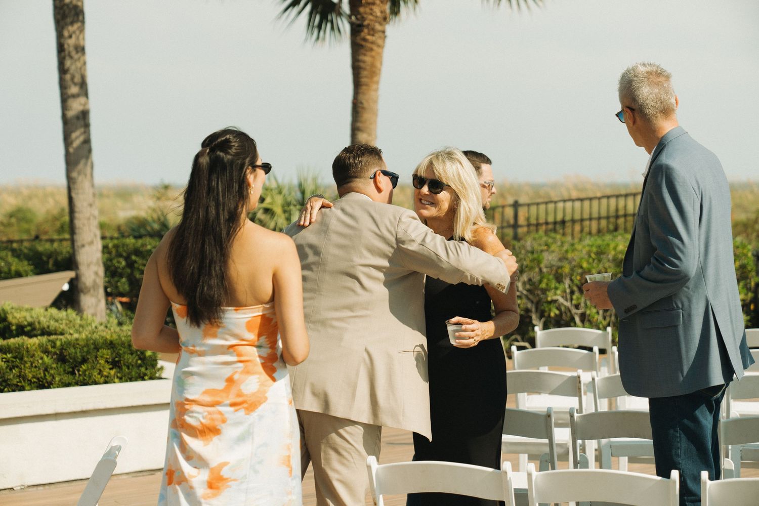 Guests mingling at an outdoor wedding ceremony with white chairs, palm trees, and a sunny coastal backdrop.