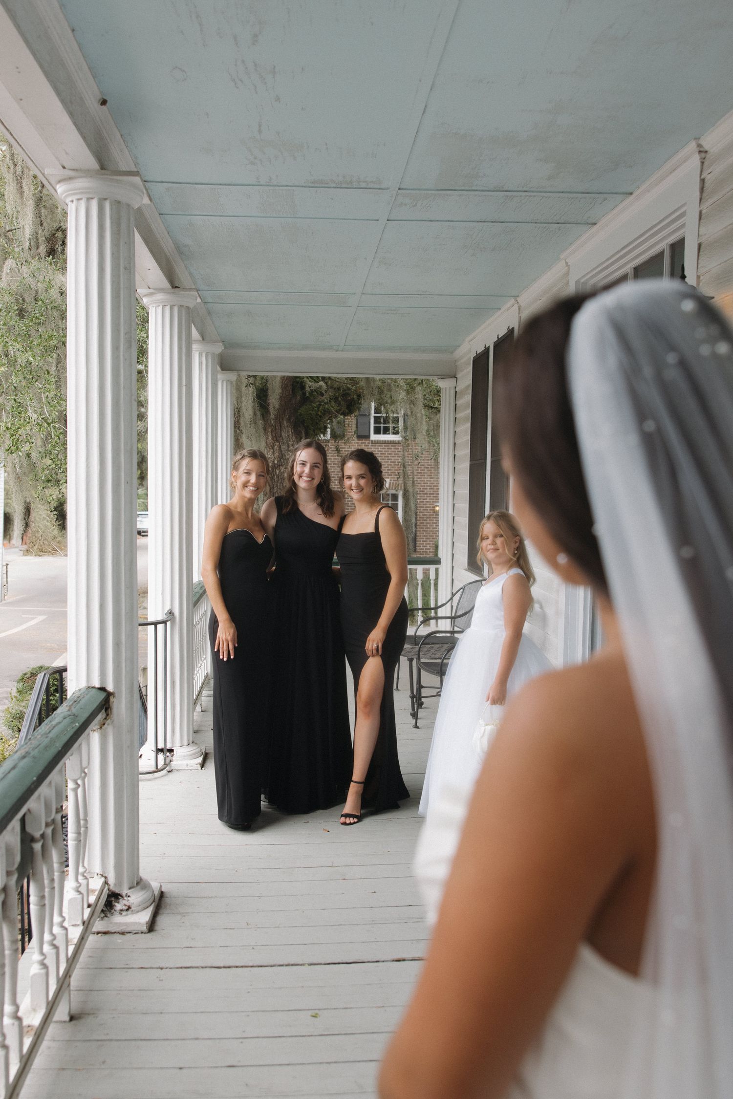 Bride in white gown and veil shares first look moment with bridesmaids in black dresses on a classic white porch.
