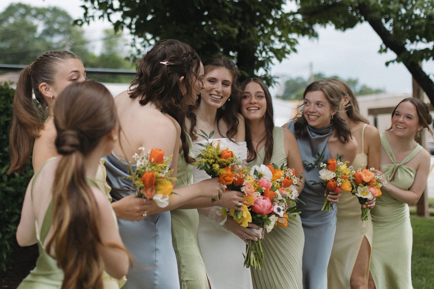 Joyful bride shares a kiss with smiling bridesmaids holding colorful bouquets at outdoor wedding.