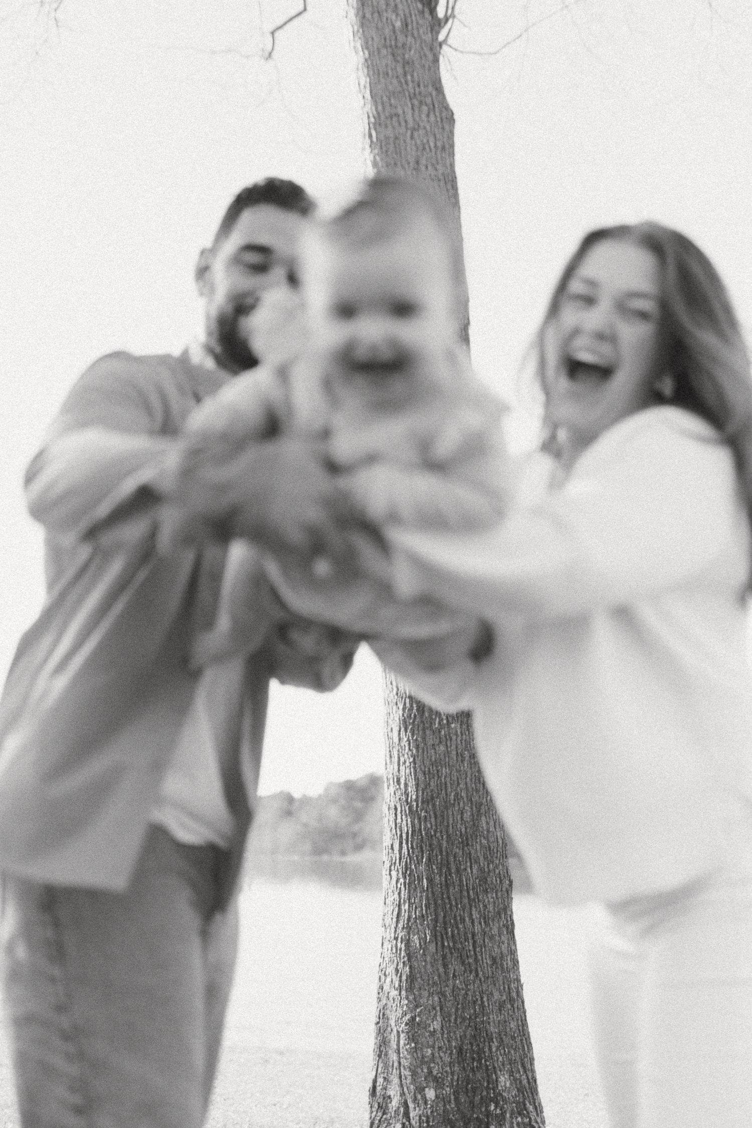 Black and white photo of laughing parents holding their smiling baby toward the camera near a tree.