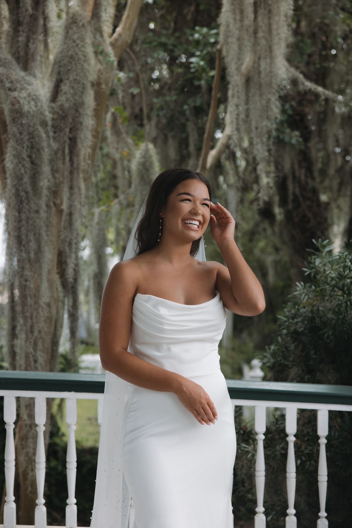 Smiling bride in white strapless gown with veil poses on balcony surrounded by moss-draped trees.