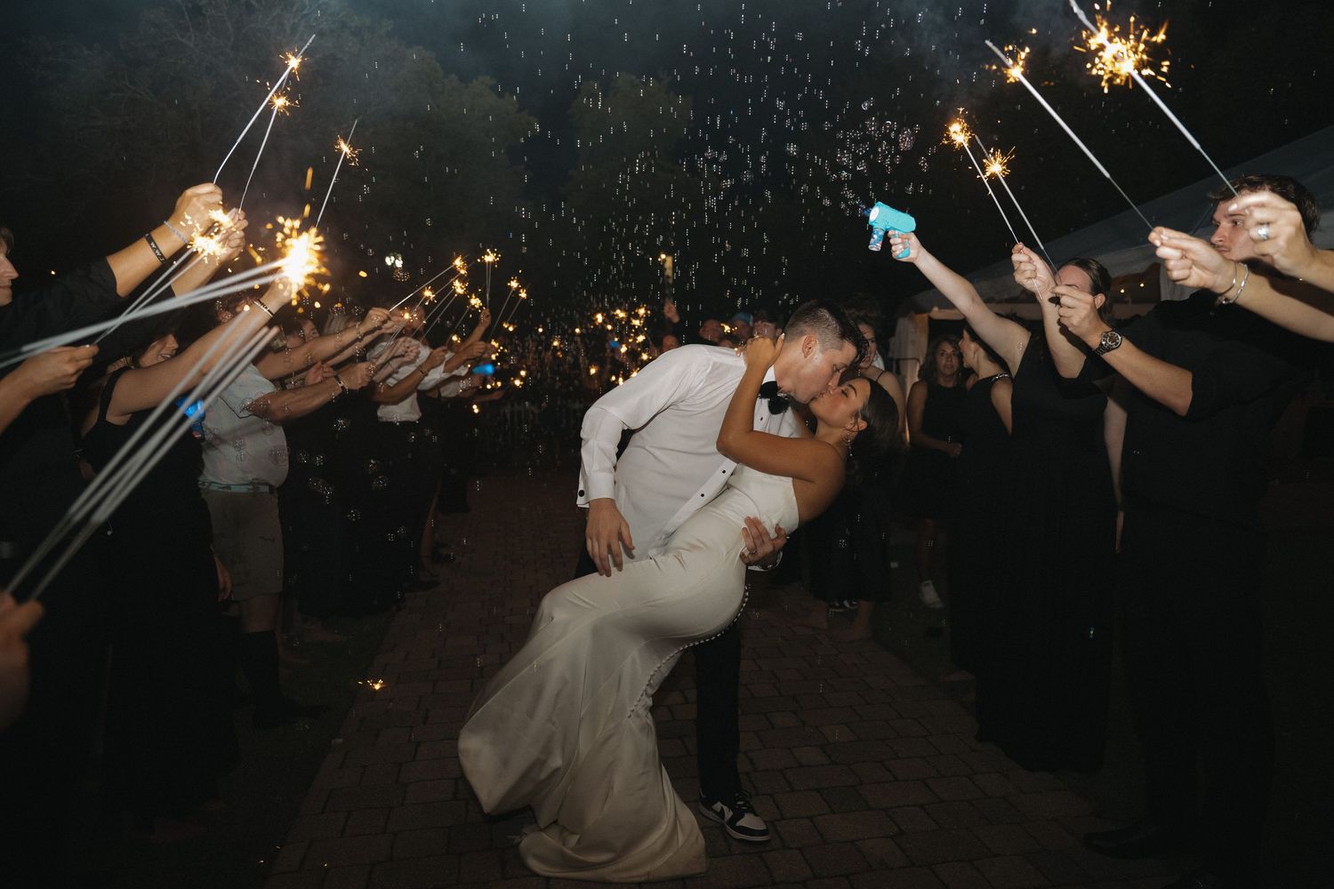 Bride and groom sharing romantic dip kiss surrounded by sparklers during nighttime wedding sendoff.