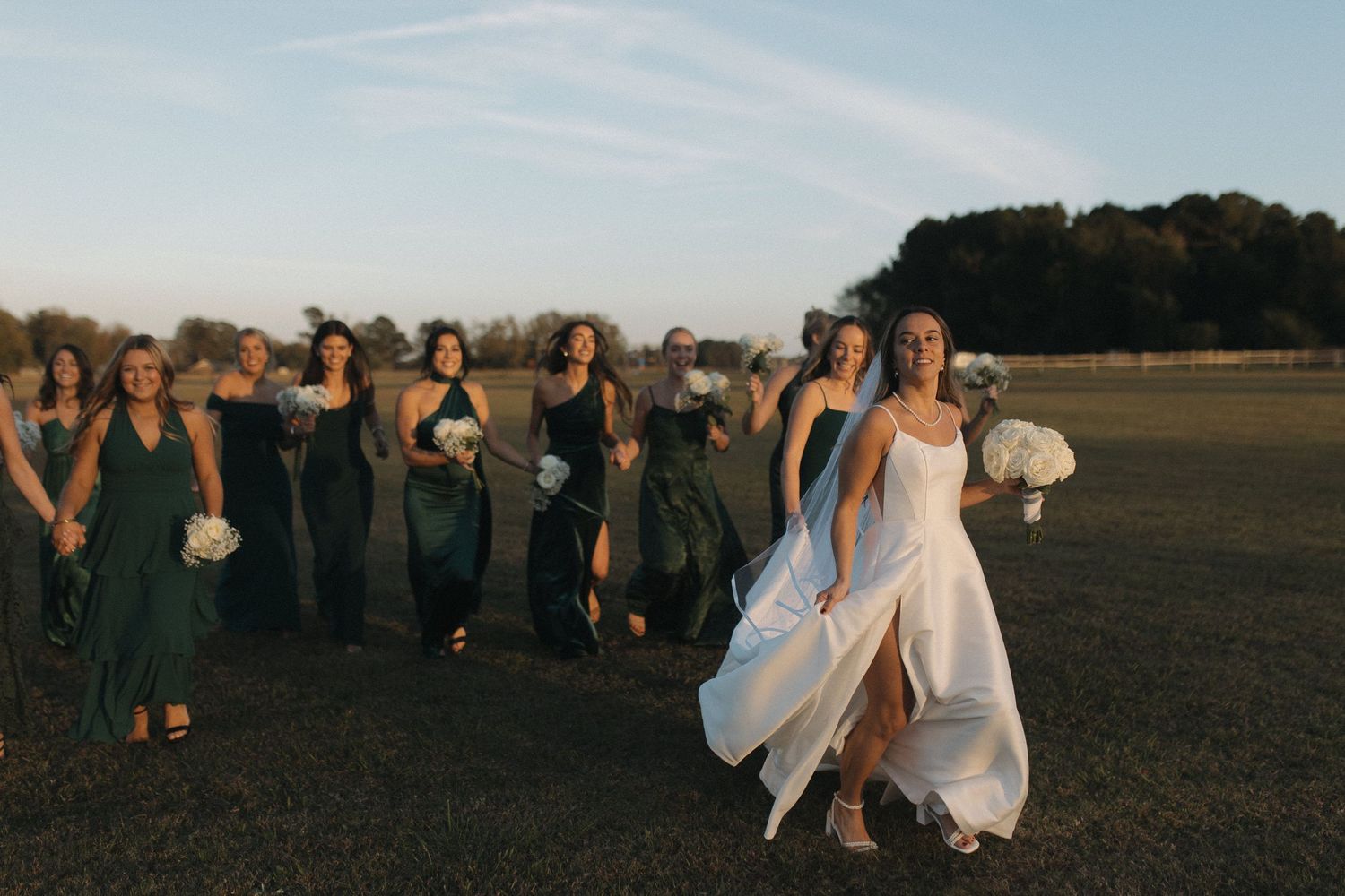 Bride in white gown walks with bridesmaids in dark green dresses across a sunlit field during golden hour.