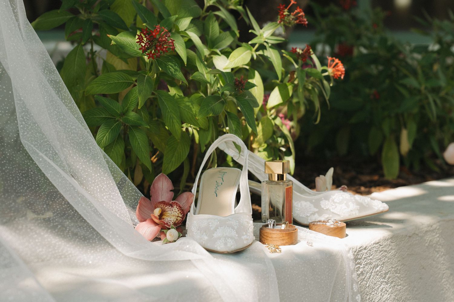 Elegant wedding flat lay with white bridal shoe, candle, rings, and floral accents on white fabric outdoors.