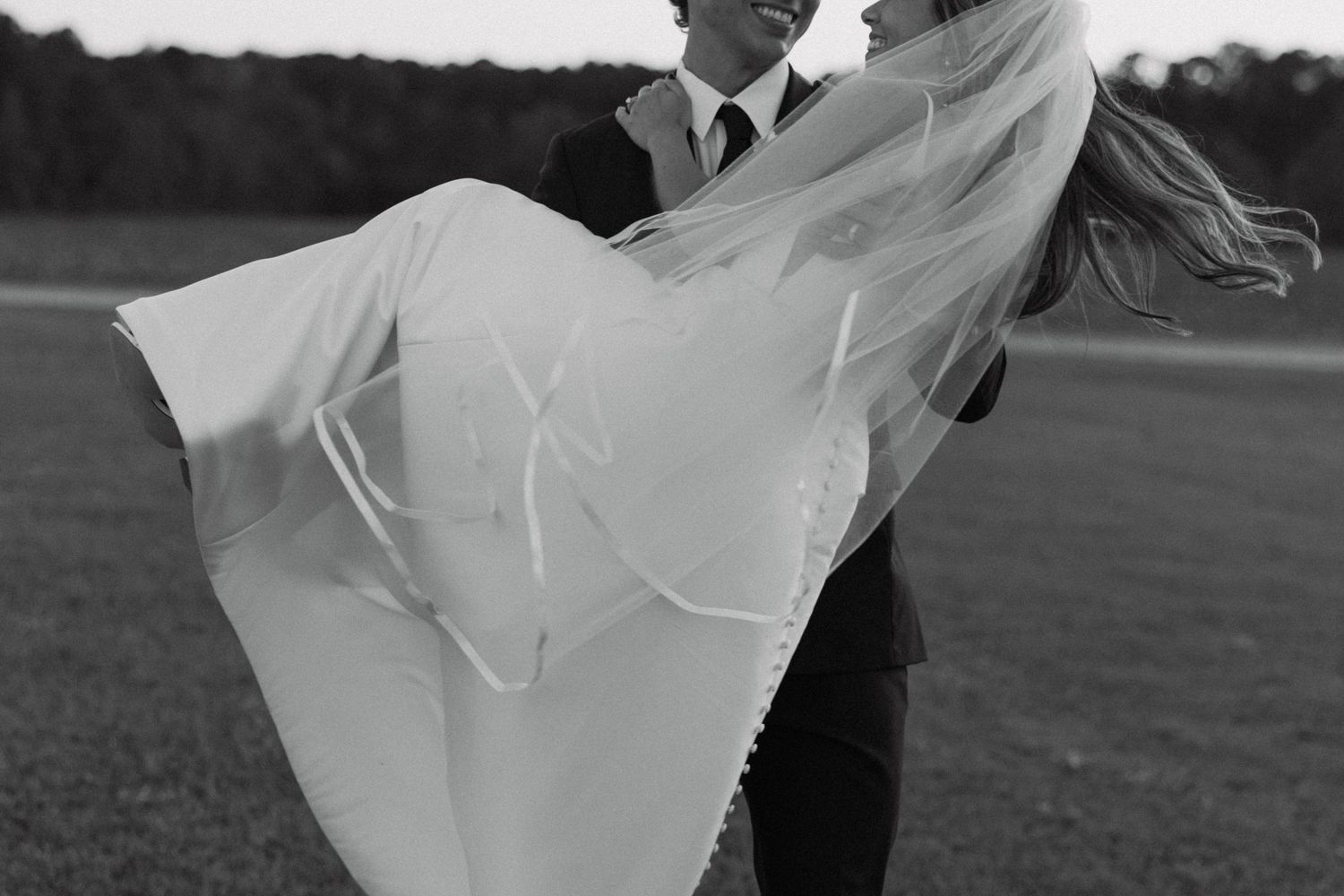 Groom lifting bride in wedding dress with flowing veil on open field.