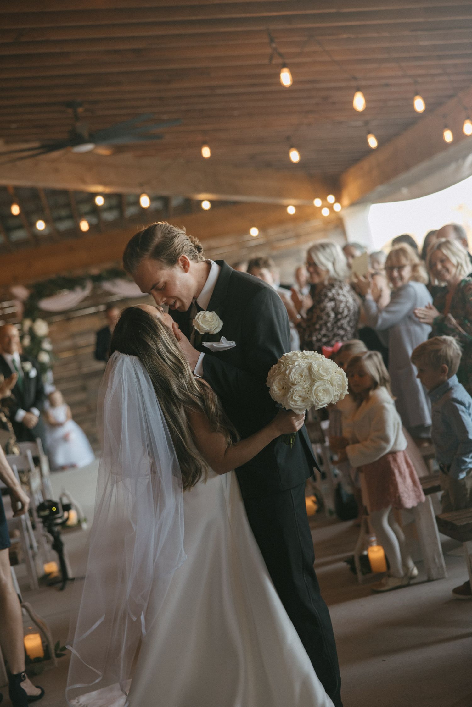 Bride and groom sharing first kiss at altar in rustic barn venue with string lights and wedding guests watching.