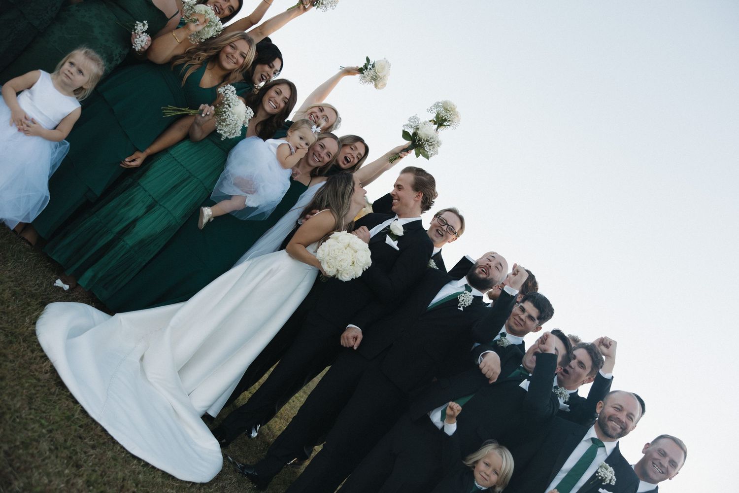 Wedding party poses together outdoors, bridesmaids in emerald green gowns and groomsmen in black suits flanking the bride and groom.