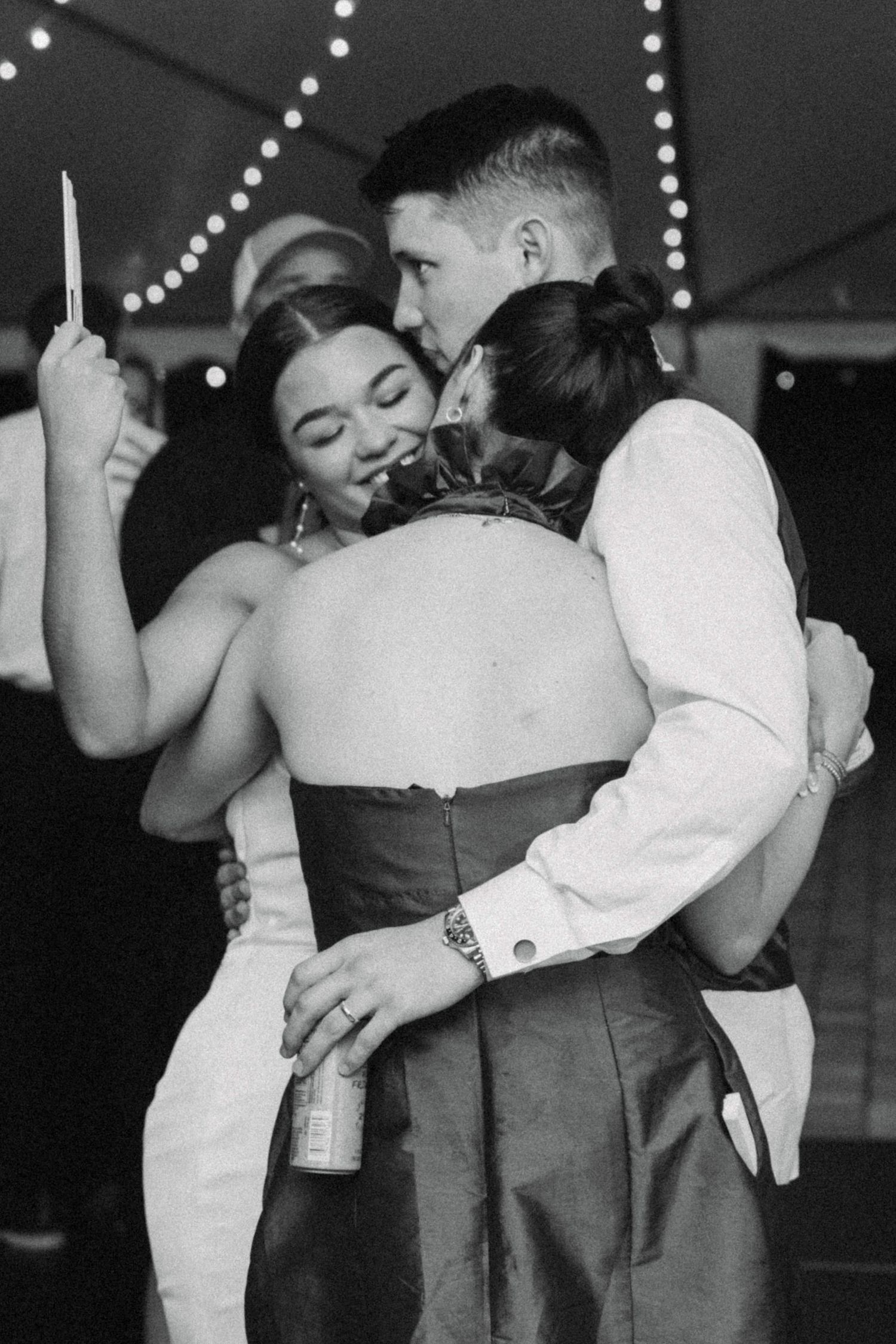 Black and white photo of bride and groom hugging friend on dance floor at wedding reception.