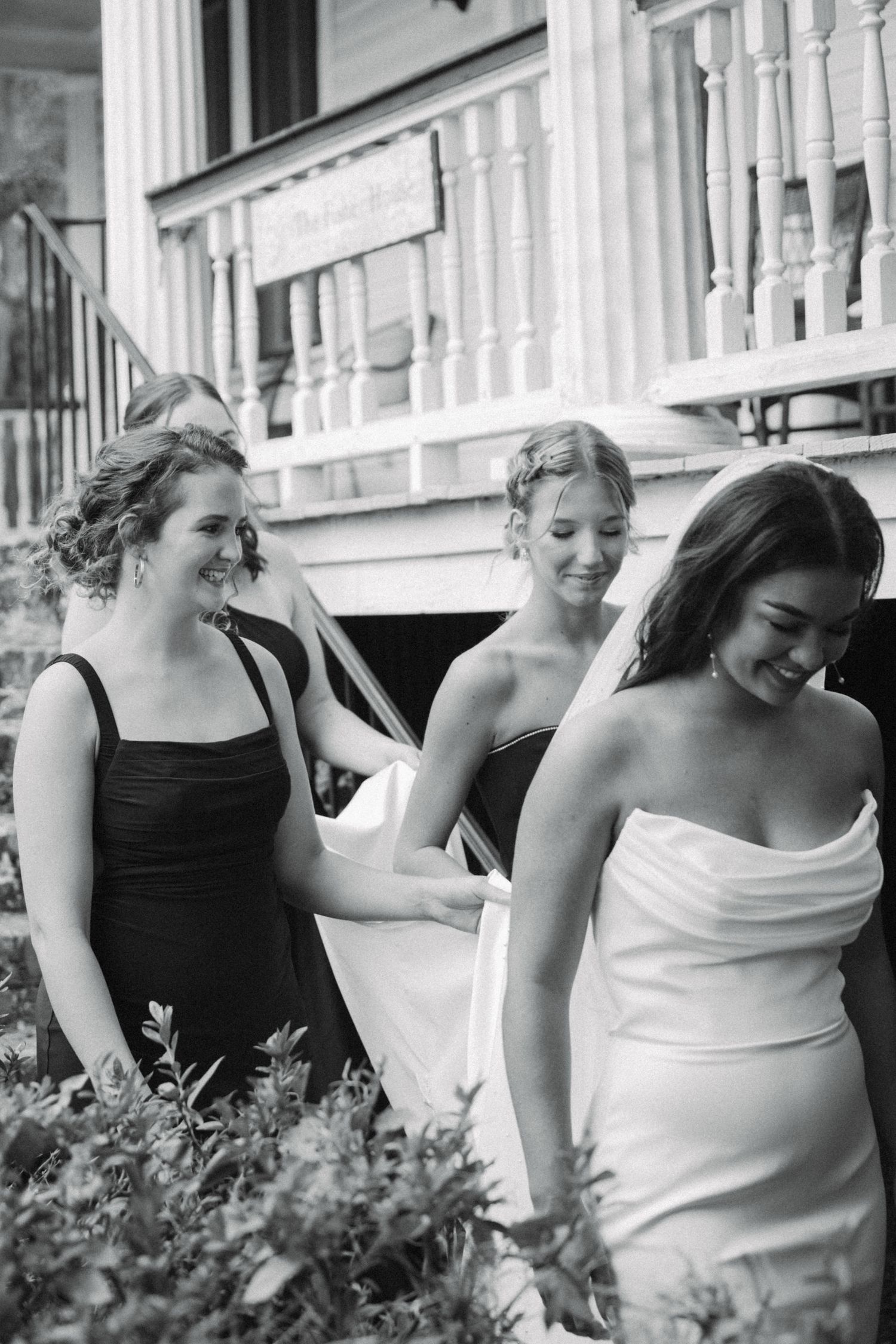 Black and white photo of a bride in white gown with bridesmaids in dark dresses walking near a porch.