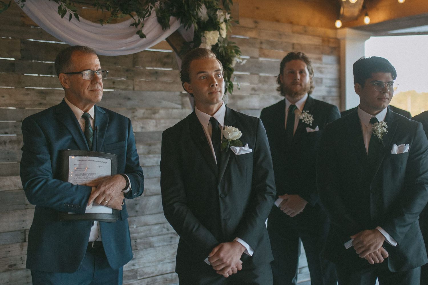 Four men in navy suits stand at a wedding ceremony altar, with the groom smiling and groomsmen beside him.