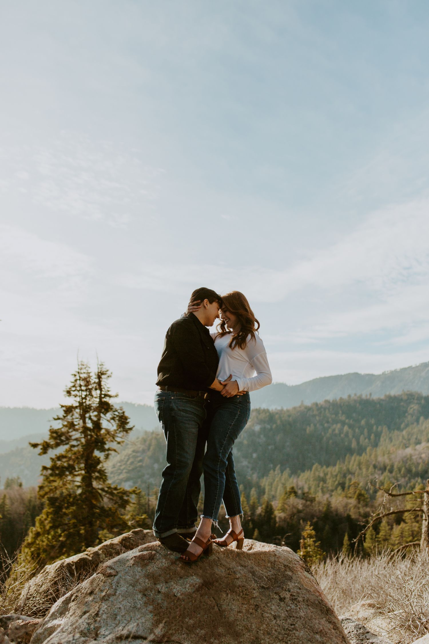 Romantic couple standing on a mountain rock, nearly kissing with scenic forest and sky backdrop.