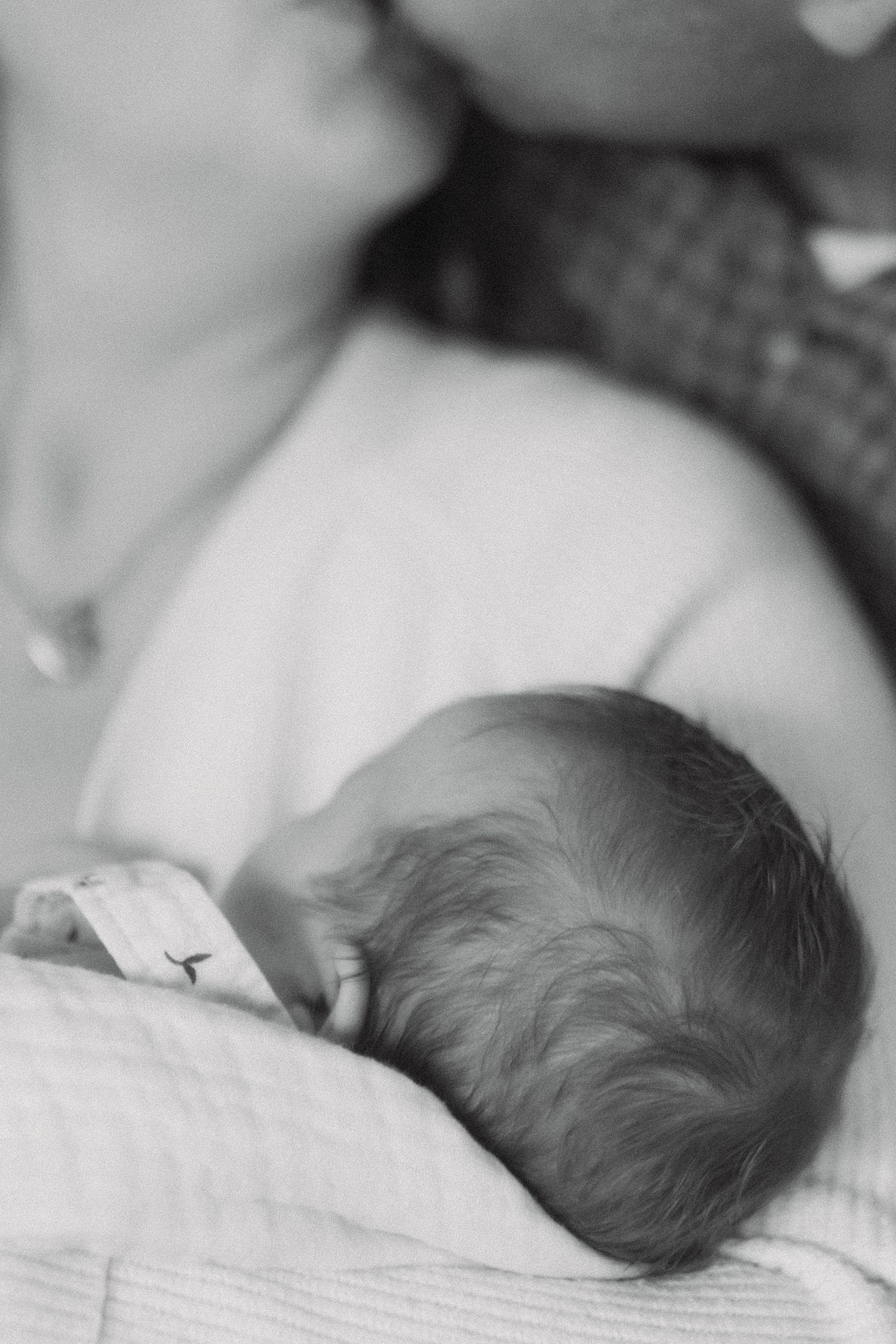Black and white close-up of newborn baby resting on mothers chest with parents nearby.