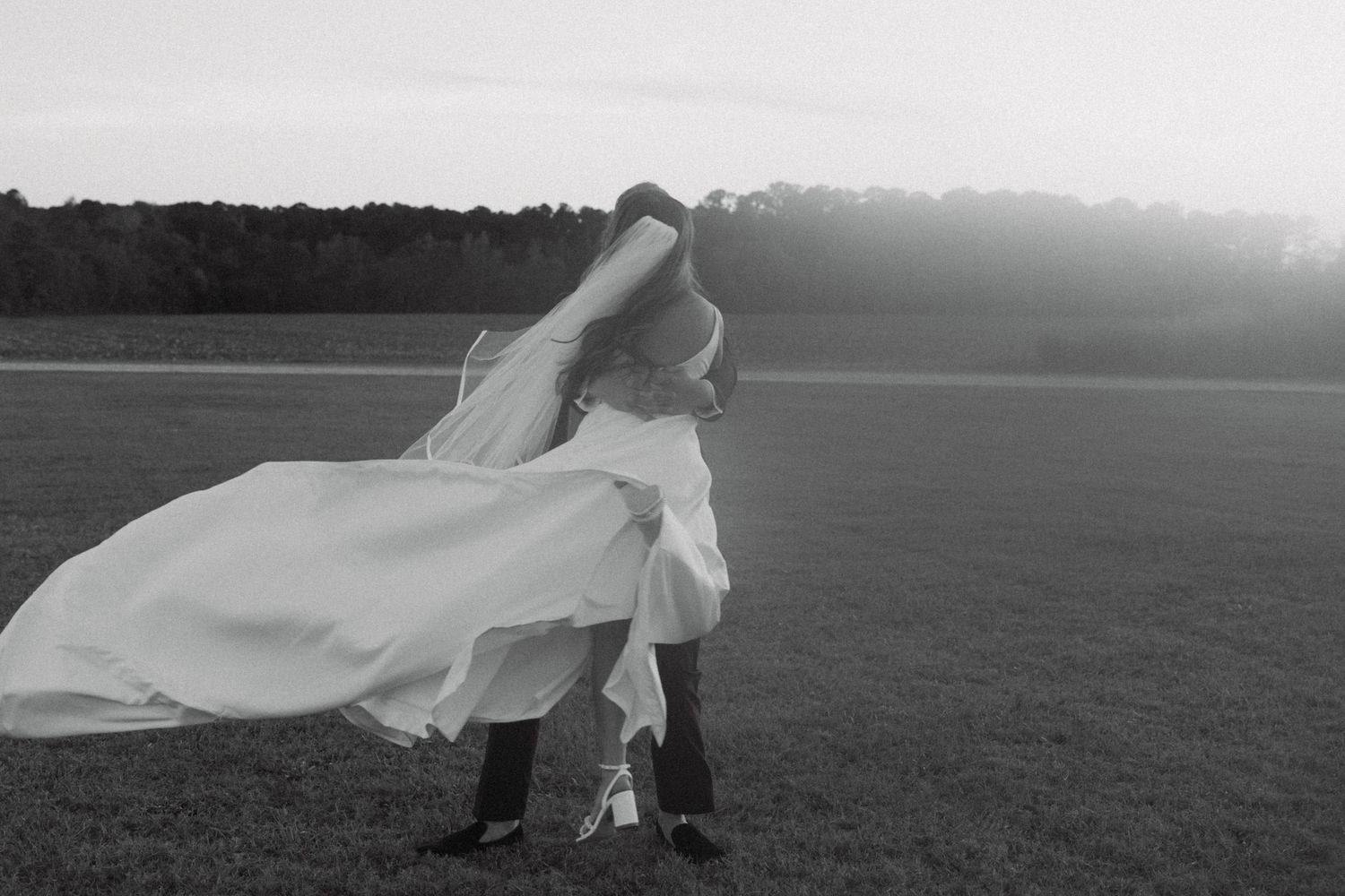 Black and white photo of bride and groom sharing a romantic dip kiss in an open field at sunset.