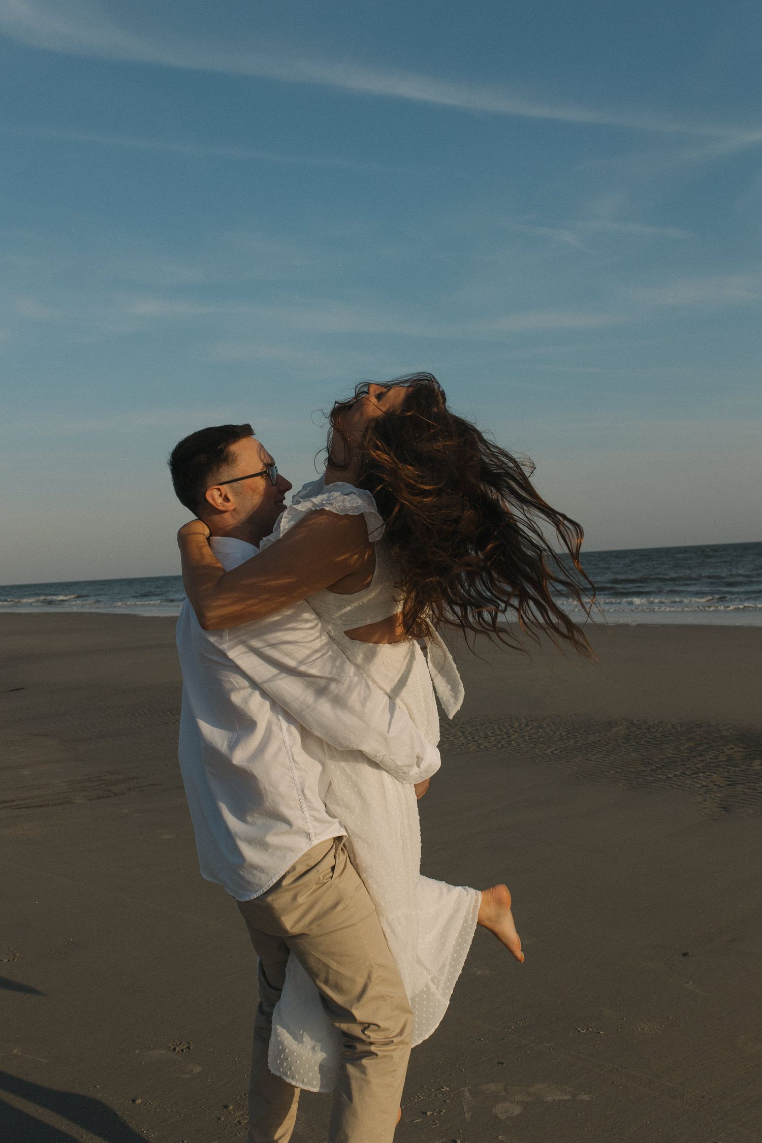 Man joyfully lifts woman on a sandy beach at golden hour by the ocean.