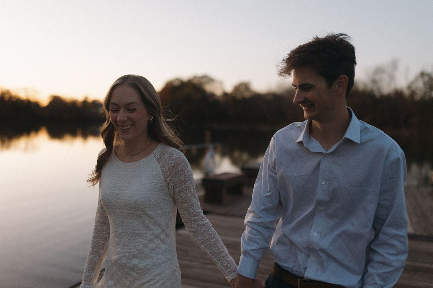 Happy couple holding hands and walking on a lakeside dock at dusk, smiling together.