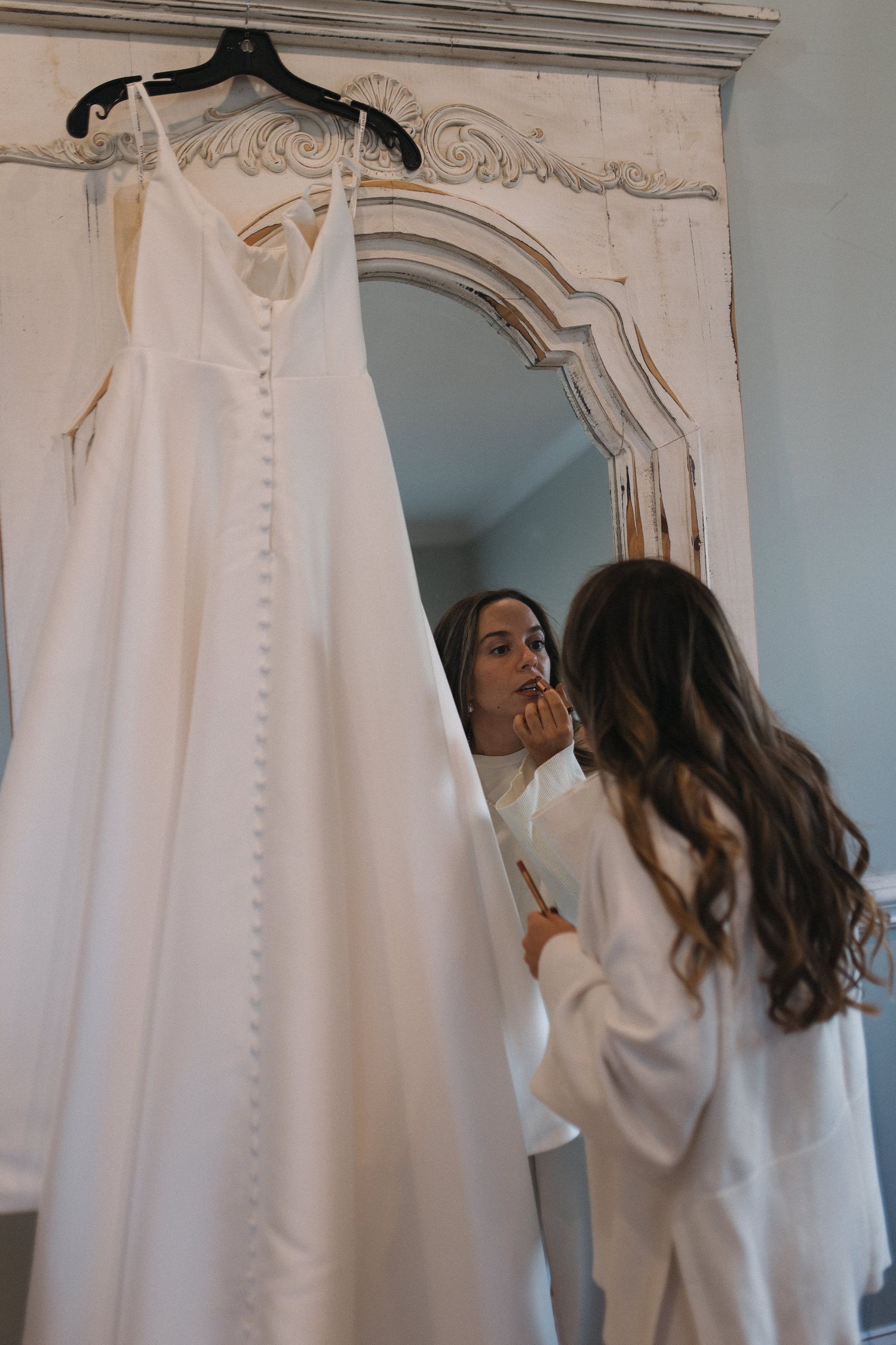 Bride applying lipstick in ornate vintage mirror beside hanging white button-back wedding gown while wearing silk robe.