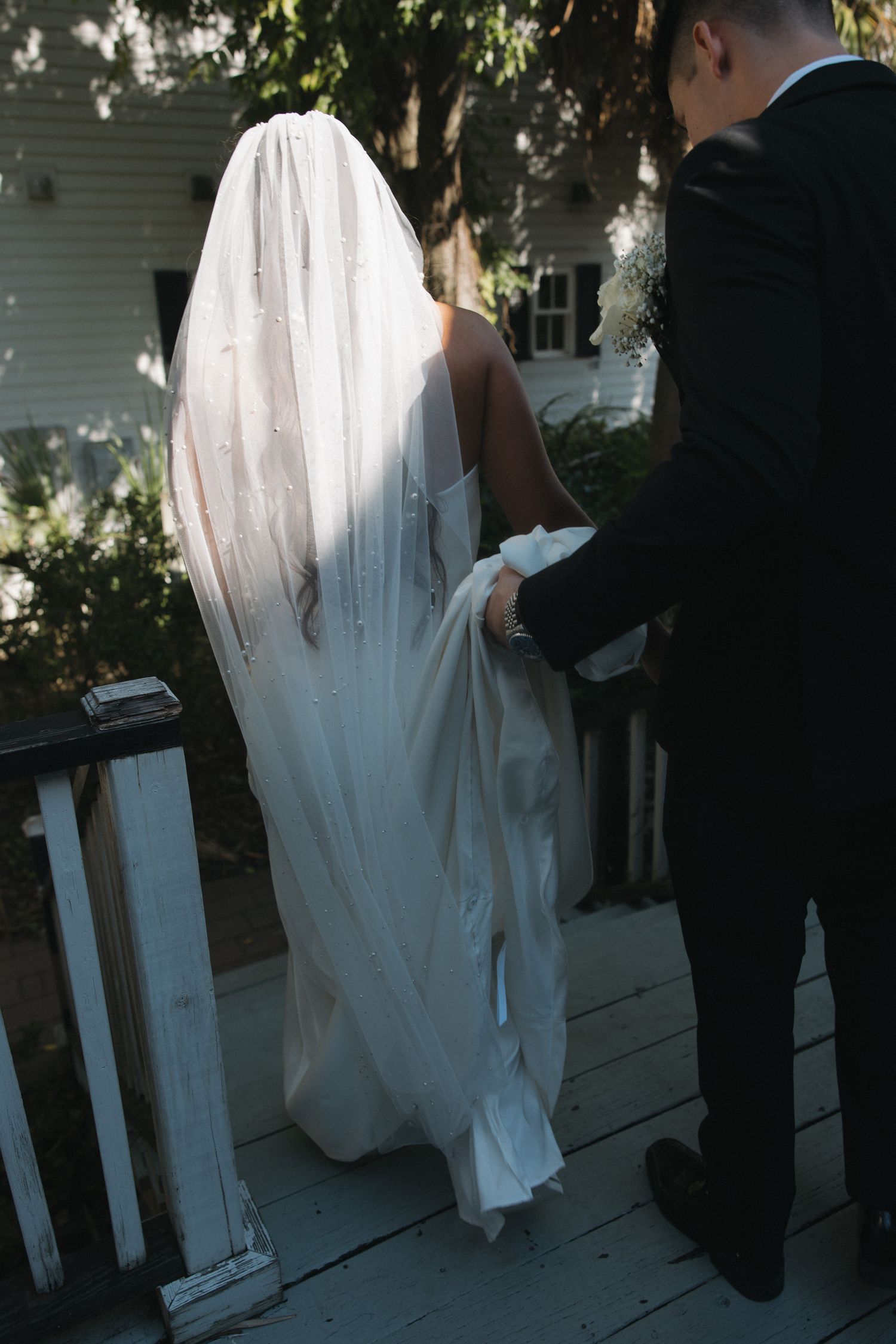 Groom in black tuxedo lifts brides white veil and train as they walk up wooden steps at their wedding venue.