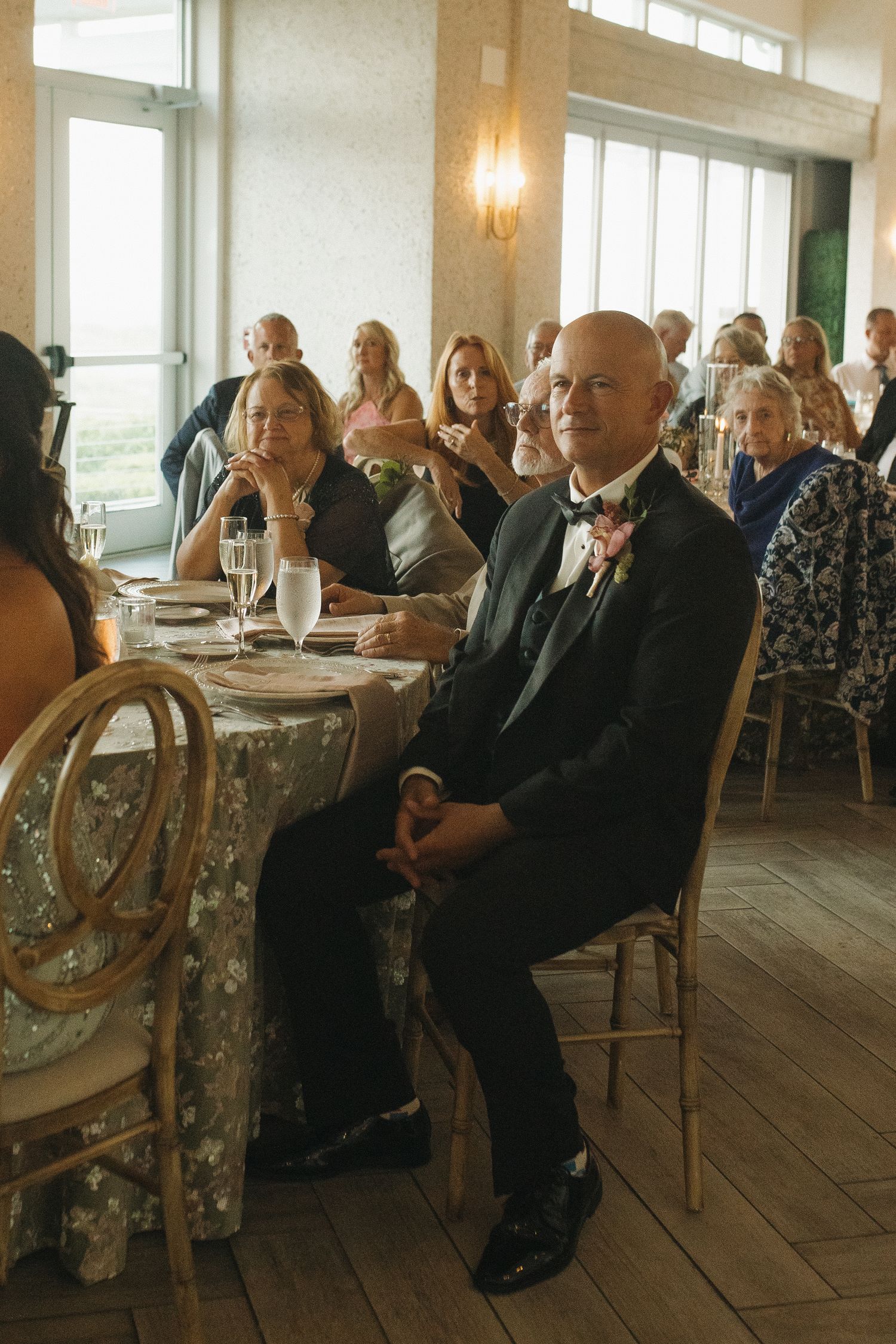 Guests seated at elegantly decorated tables during a wedding reception, with a bald man in a tuxedo smiling in the foreground.