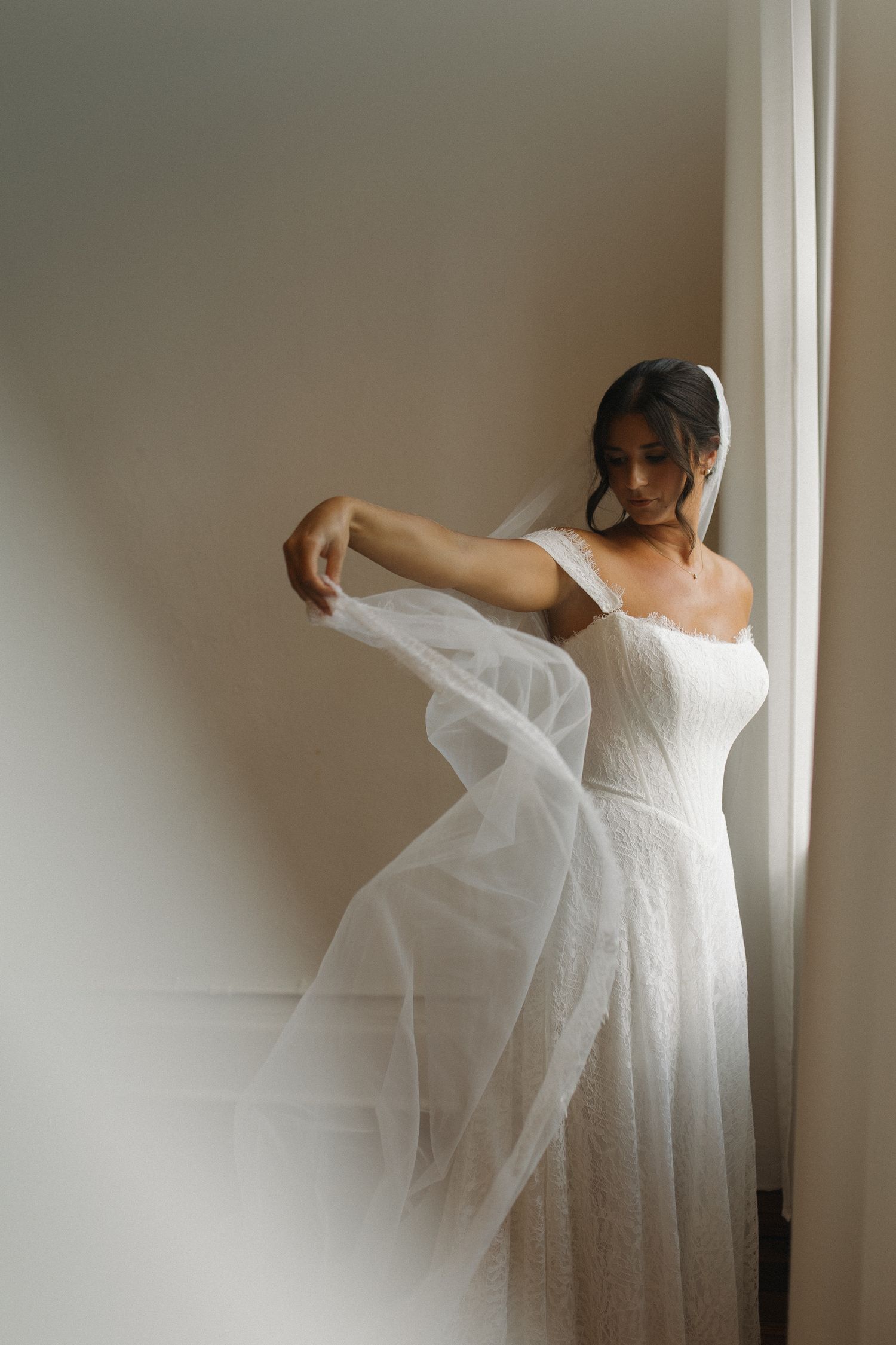 Bride in off-shoulder white lace wedding gown holding flowing tulle veil near window in soft natural light.