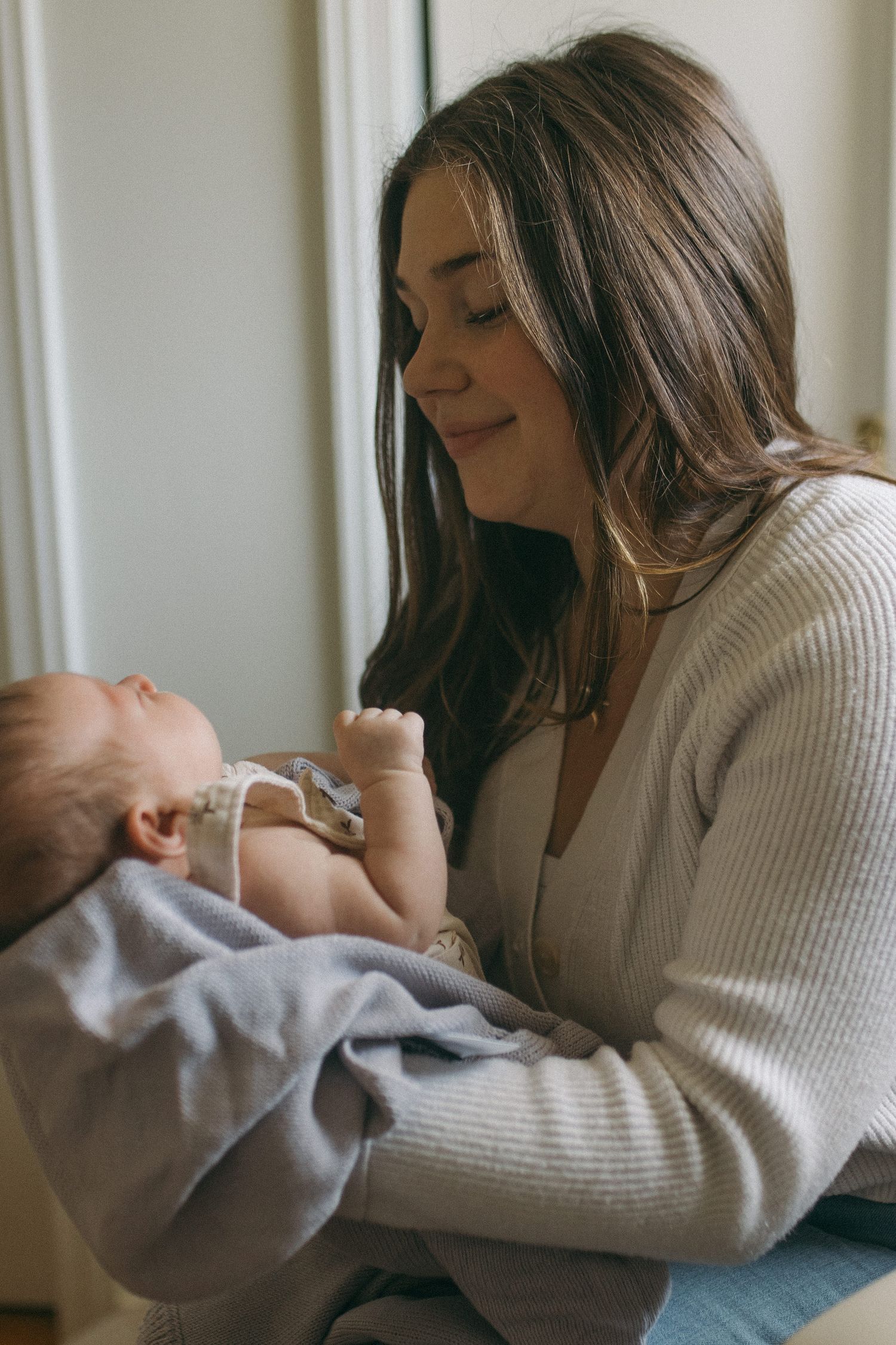 Young mother in cream cardigan smiling tenderly at her newborn baby by window.