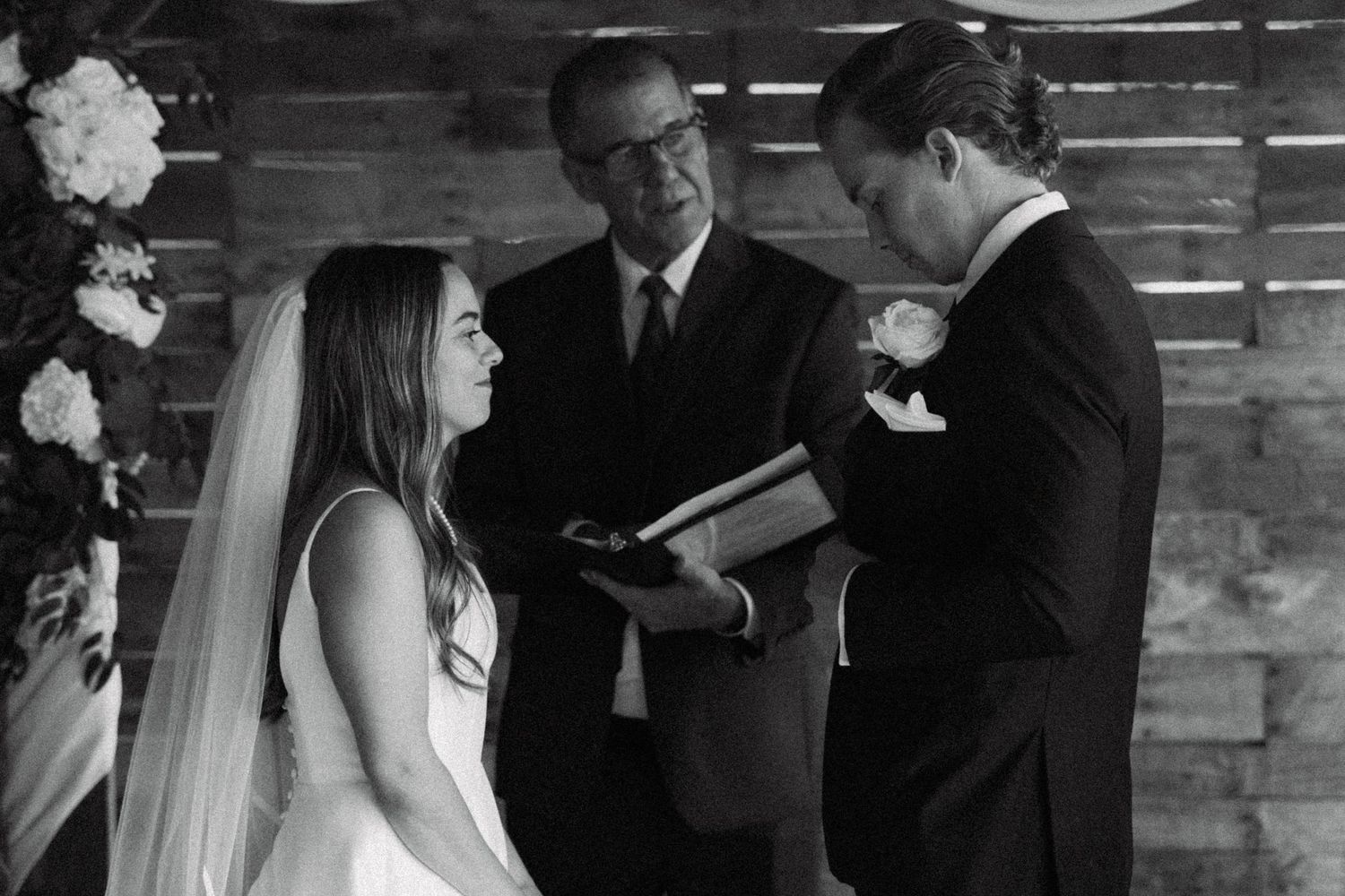 Black and white wedding ceremony photo of bride and groom exchanging vows before an officiant indoors.