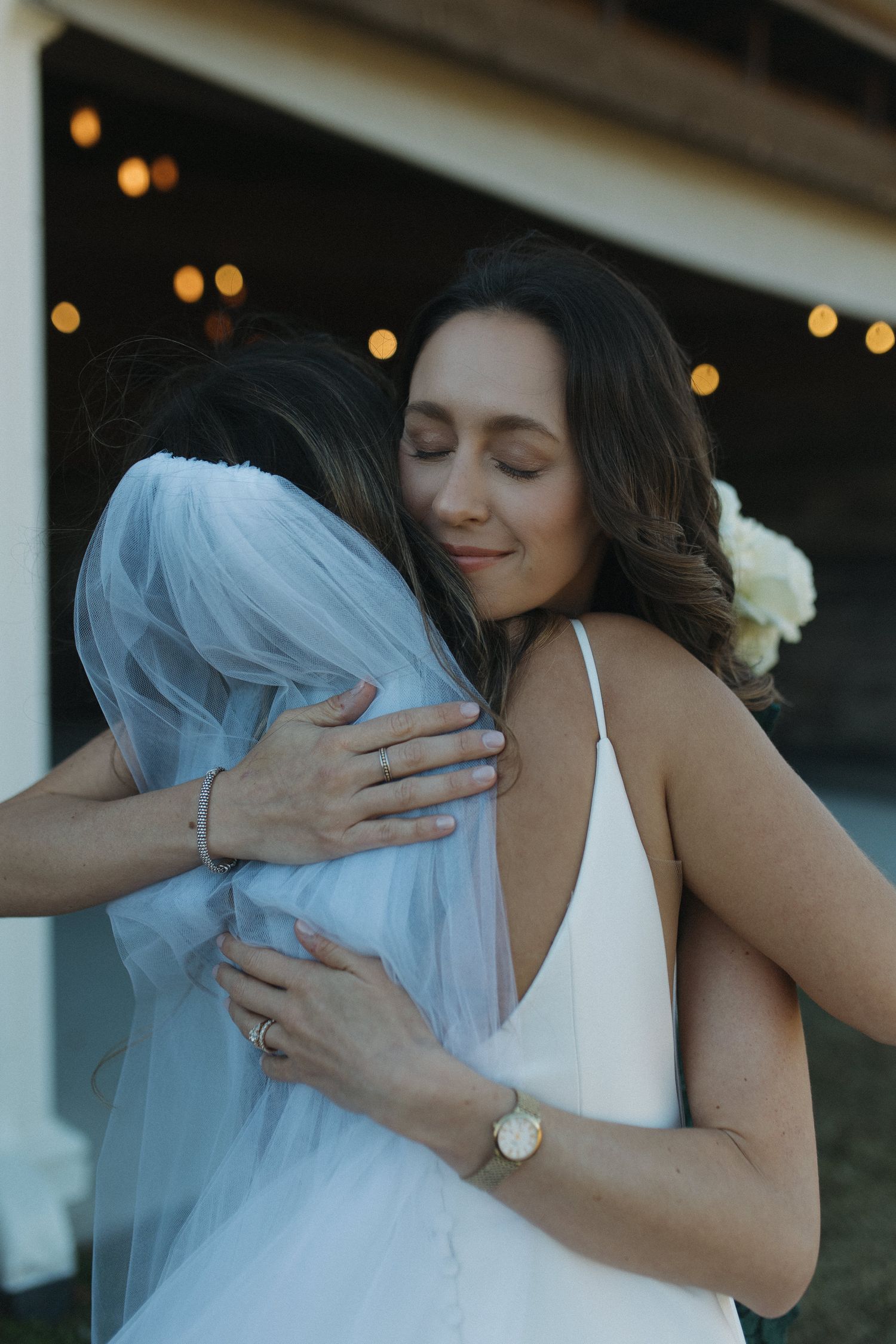 Two brides in white gowns share a heartfelt embrace outdoors, with warm string lights glowing in the background.