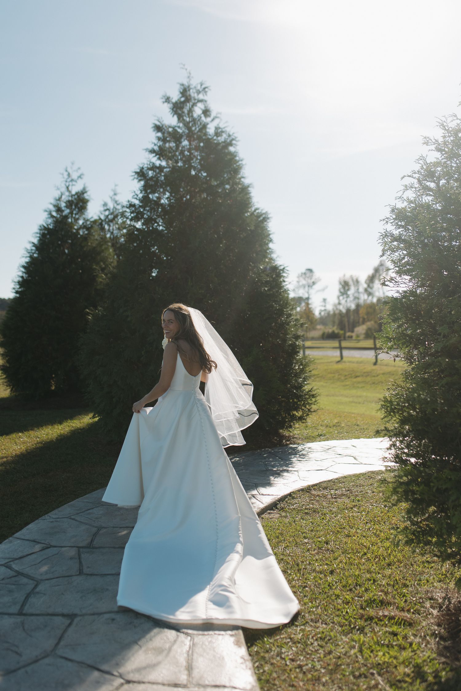 Bride in white gown and veil walks along stone path surrounded by tall evergreen trees on sunny wedding day.