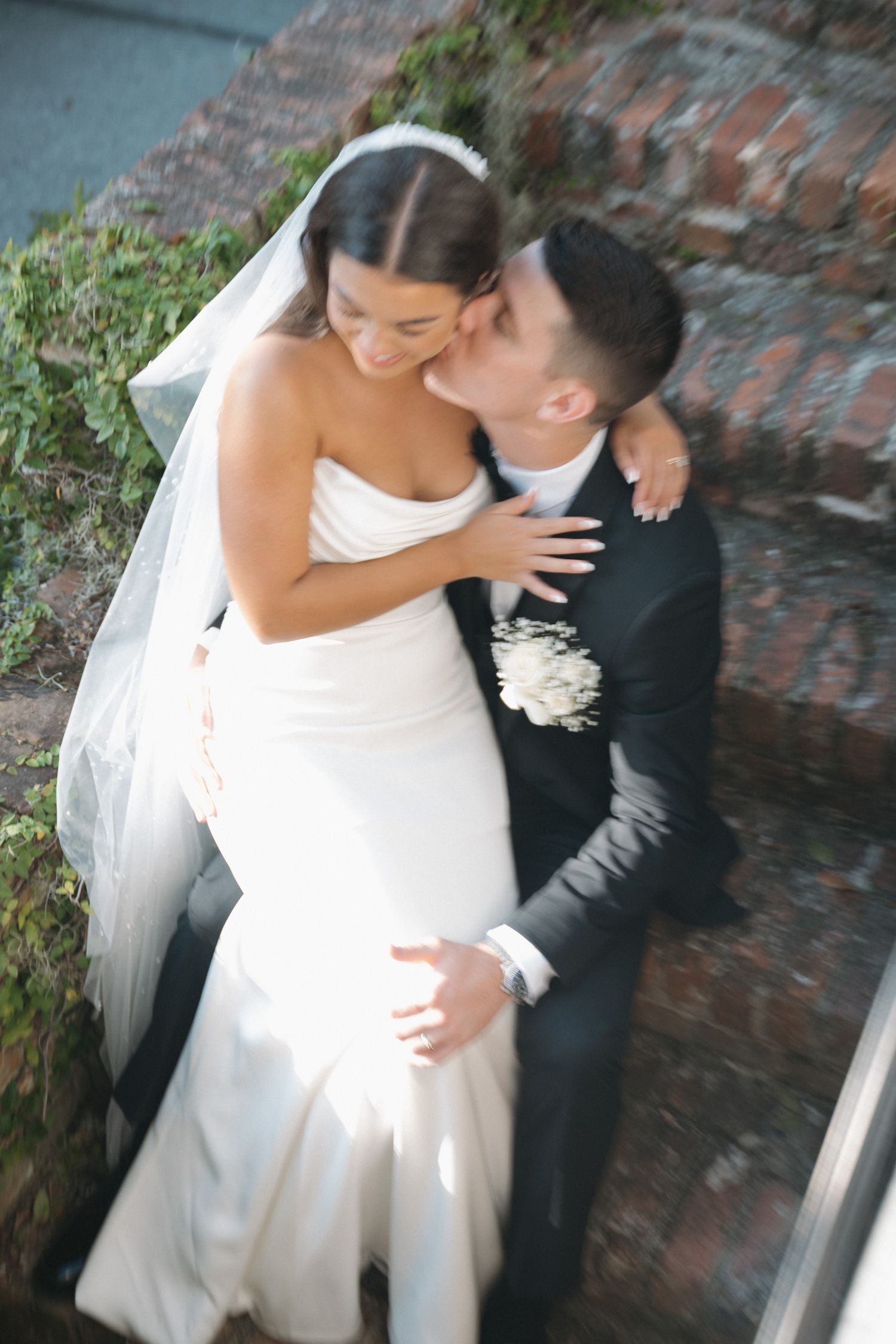 Bride and groom share a romantic kiss on outdoor stone steps, bride in white gown with veil, groom in dark suit.