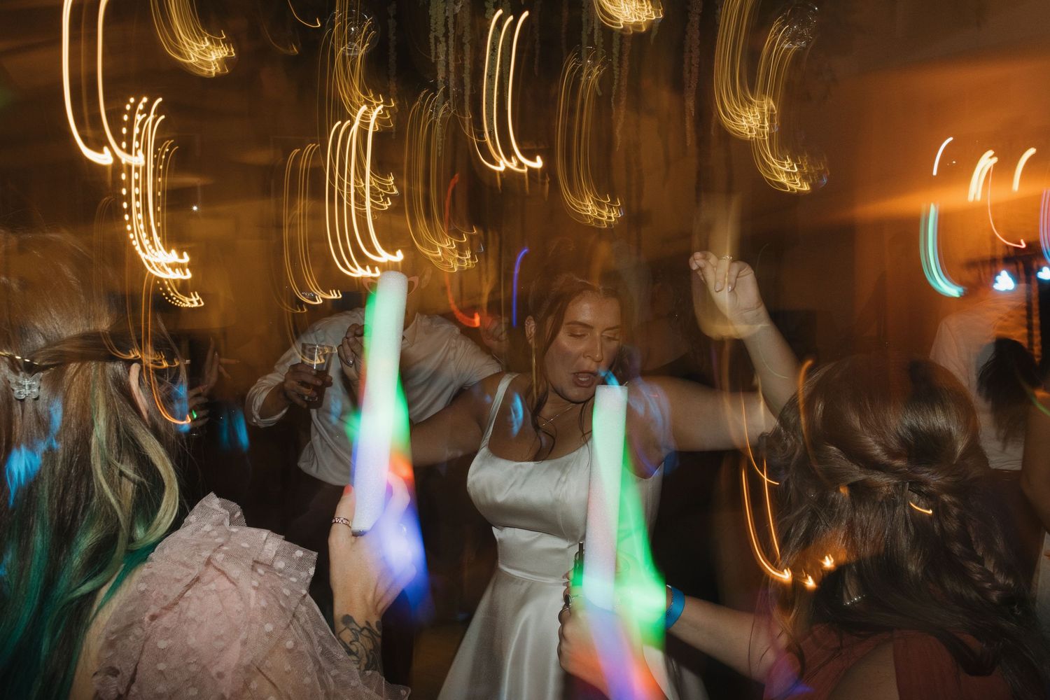 Bride dancing joyfully with guests at a wedding reception, surrounded by colorful glow sticks and streaking lights.