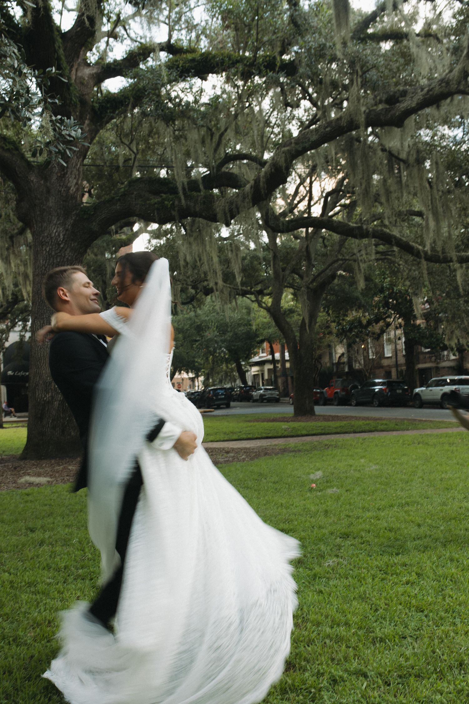 Groom lifting bride in white gown under Spanish moss oak trees in a lush green park on their wedding day.