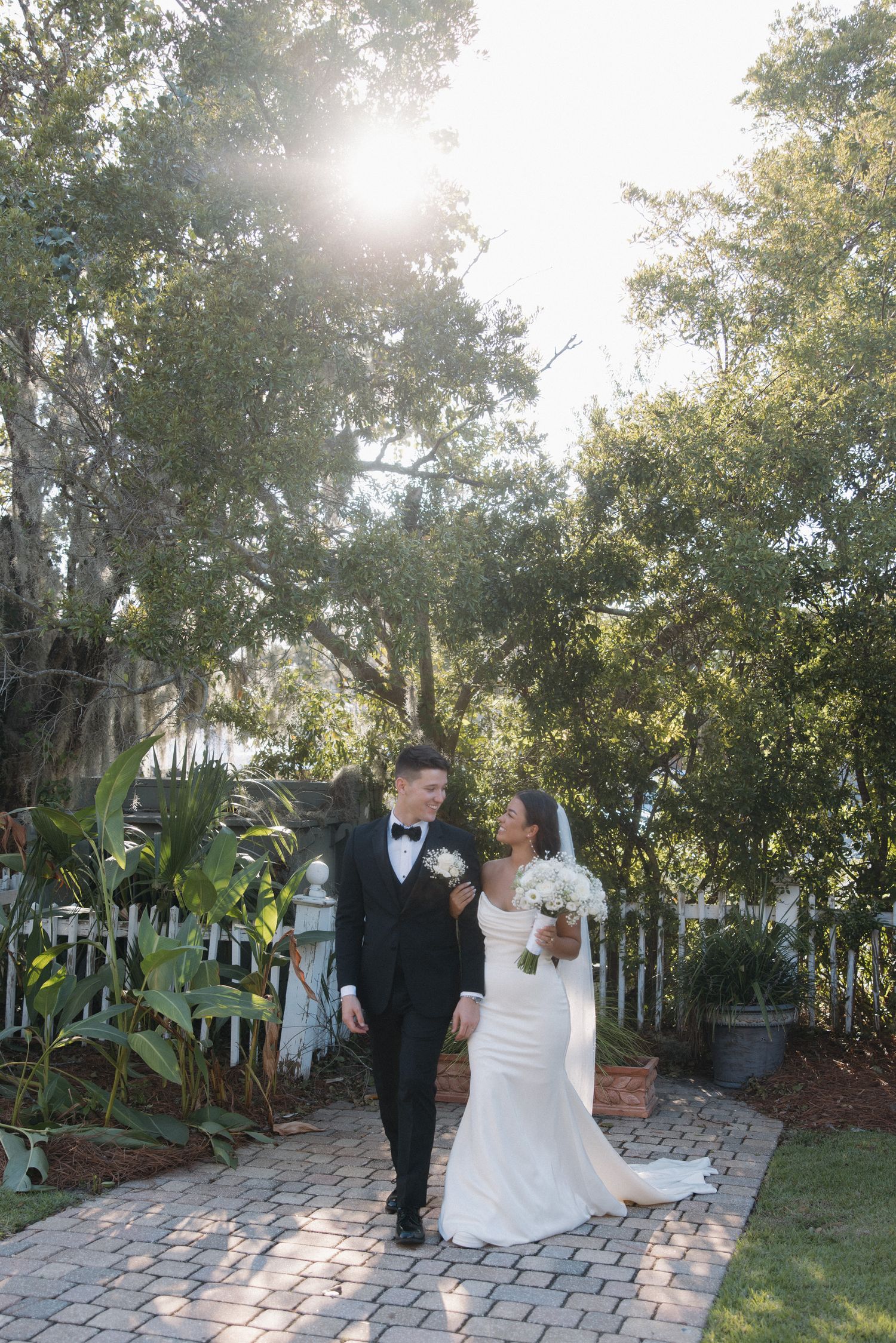 Bride and groom walk hand in hand on a sunlit brick path surrounded by lush greenery at their wedding venue.