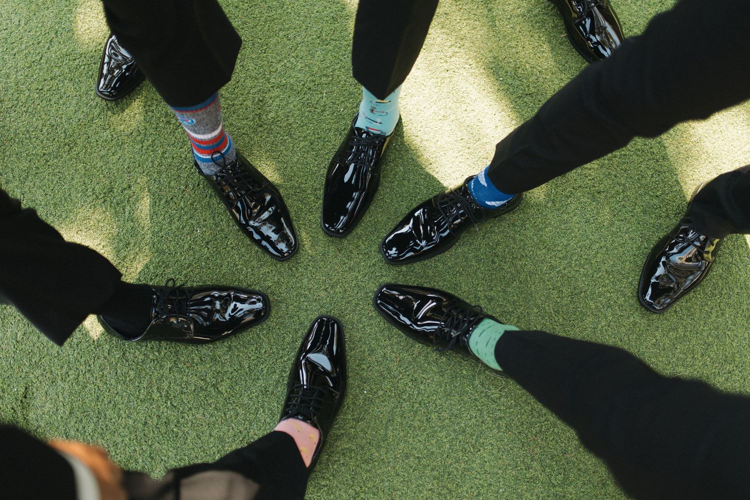 Group of men wearing matching black patent leather shoes with colorful socks arranged in a circle on green grass.