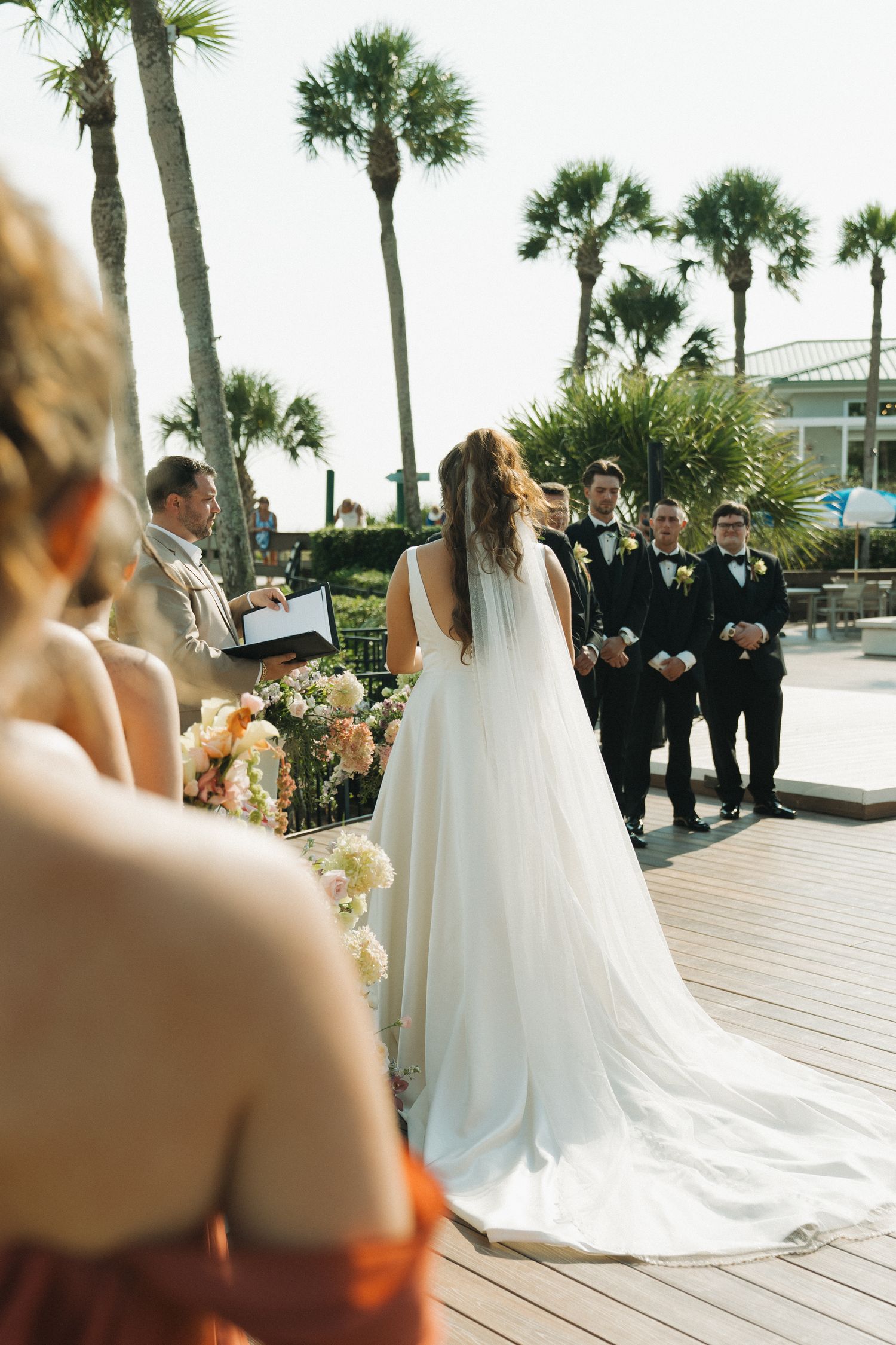 Bride in white gown stands before groom and groomsmen during outdoor wedding ceremony with palm trees.