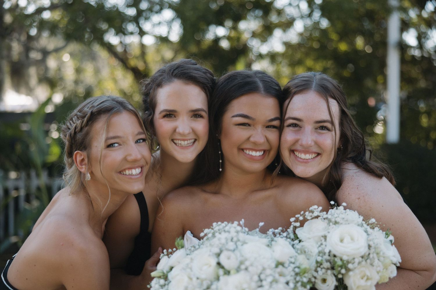 Bride holding white floral bouquet smiling with three bridesmaids in black dresses outdoors.