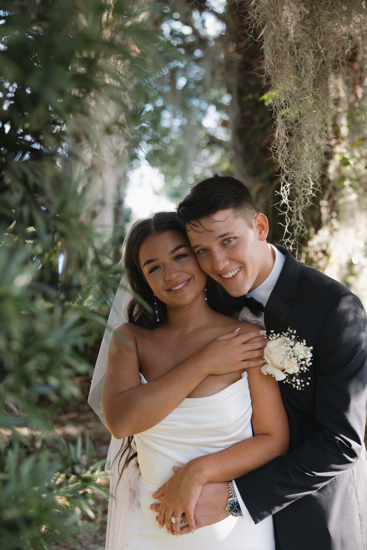 Happy bride and groom embracing and smiling under Spanish moss trees at outdoor wedding venue.