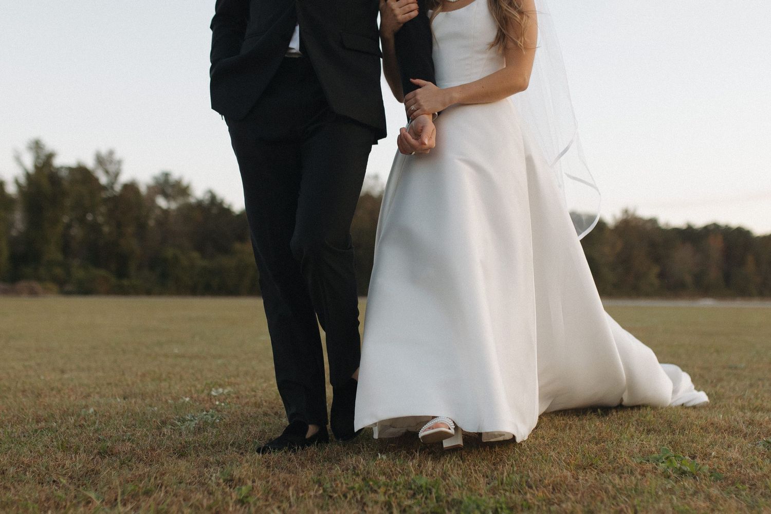 Elegant bride in white gown and groom in black suit holding hands in an open field.