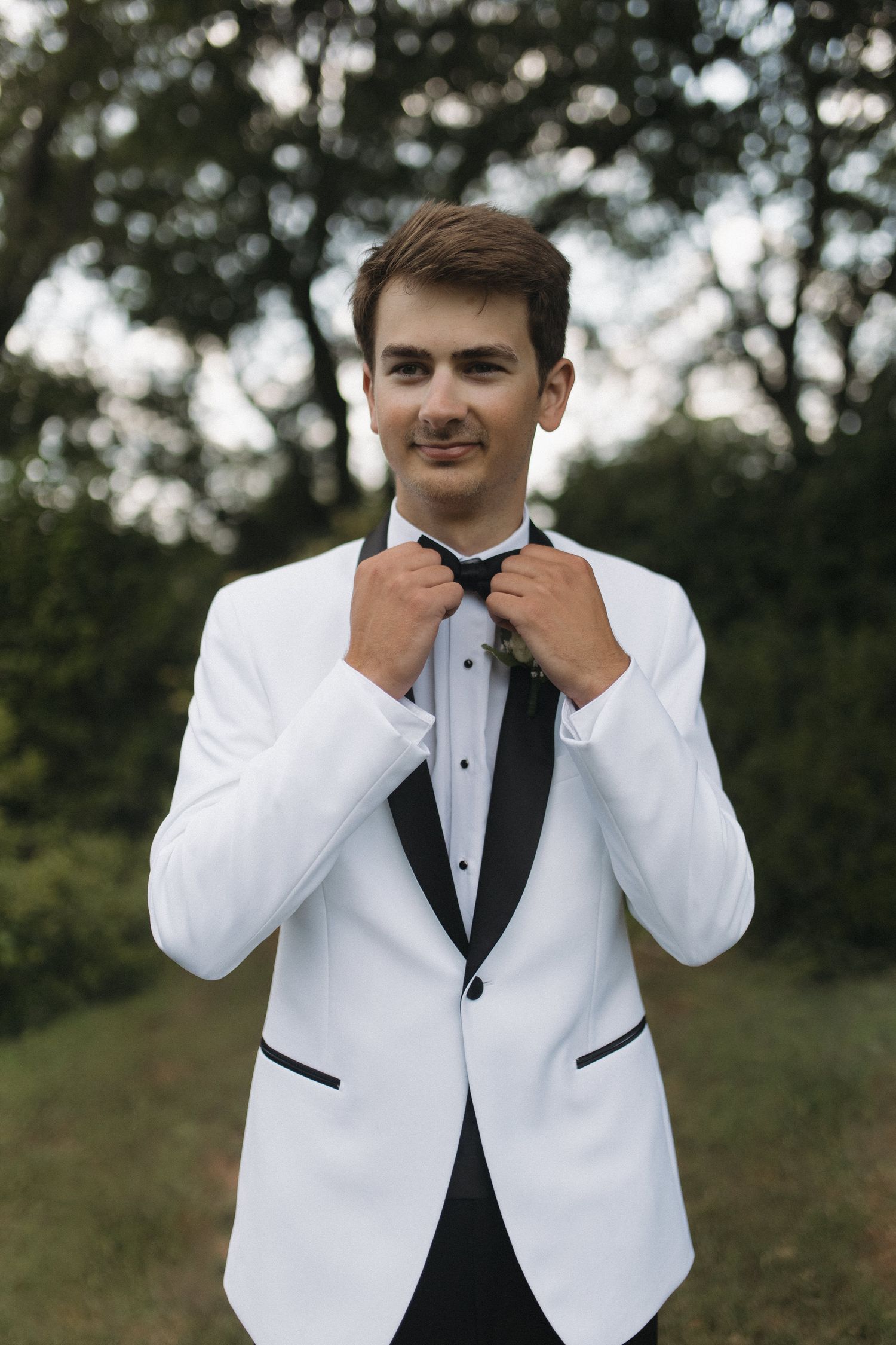 Smiling groom in a white tuxedo adjusting his black bow tie outdoors.