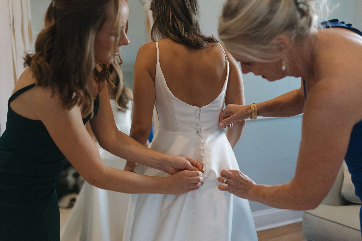 Two women helping a bride button up her white wedding dress from behind in a bright indoor setting.