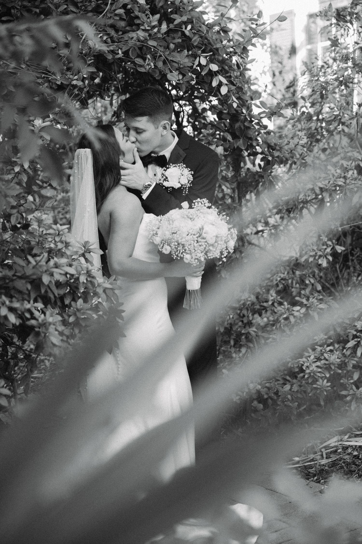 Black and white photo of newlyweds sharing a romantic kiss surrounded by lush garden greenery.