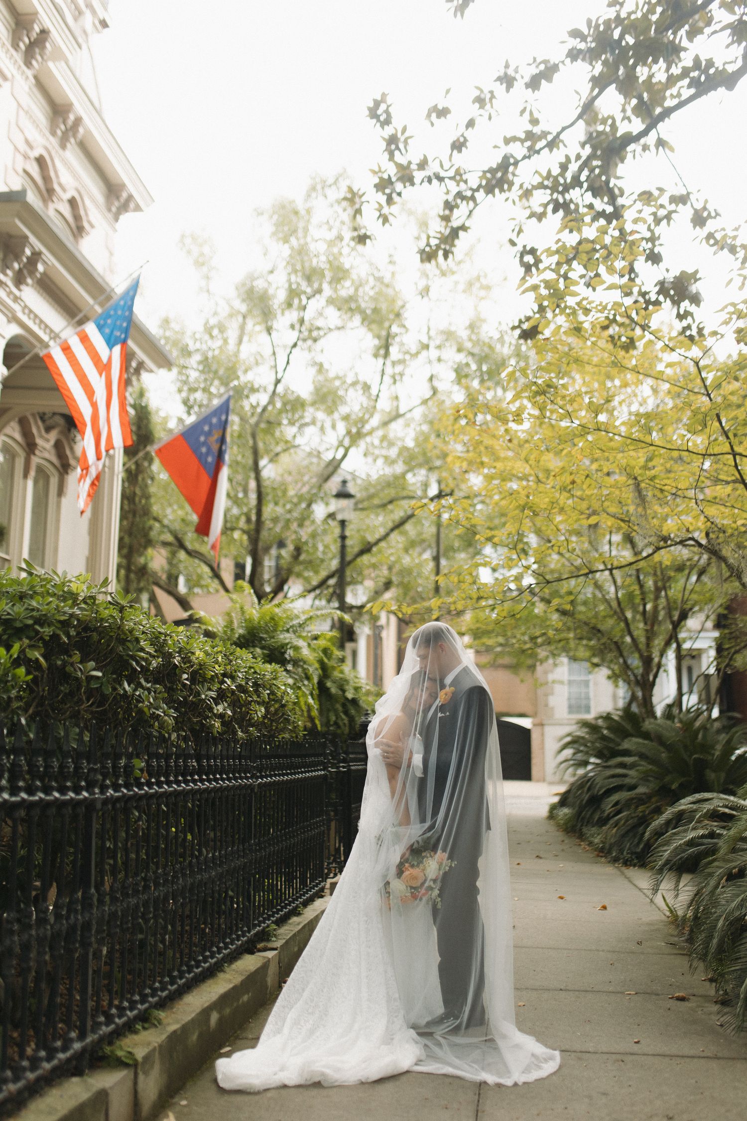 Bride and groom kissing under a flowing veil on a tree-lined sidewalk with American flags in the background.
