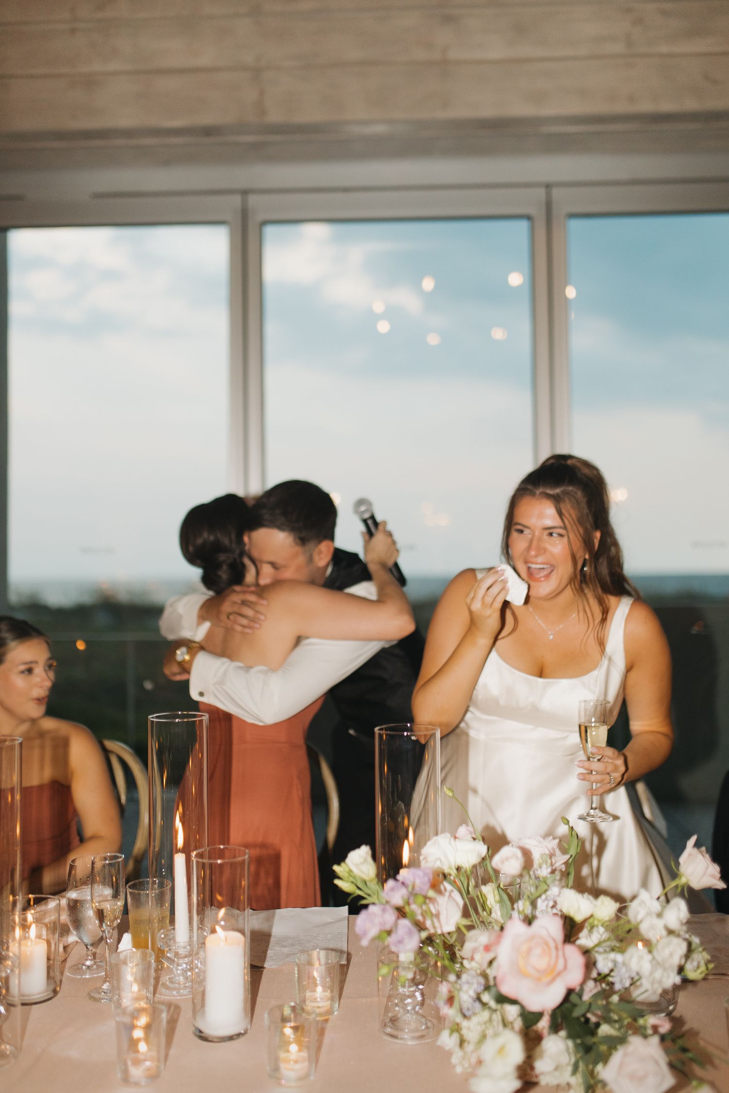 Bride wiping tears during wedding reception toast while guests hug in background near candlelit floral table.