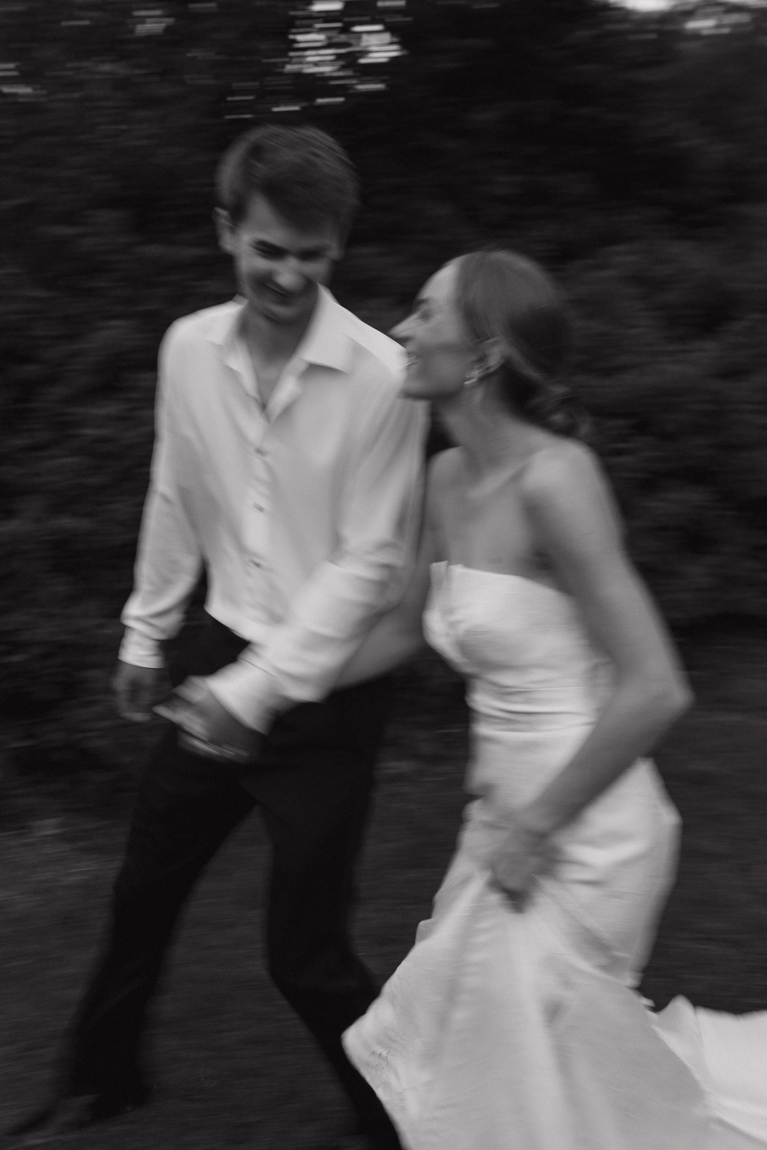 Black and white blurred motion photo of bride and groom laughing and running together outdoors.