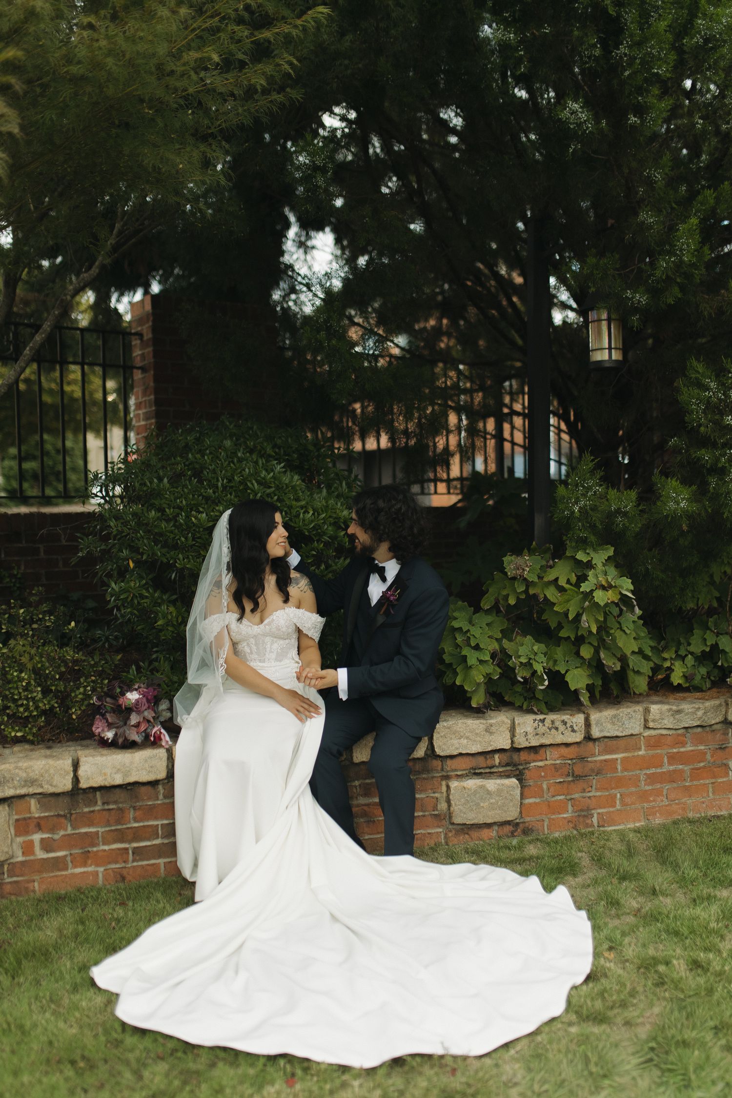 Bride in white gown with long train sits on brick wall with groom in dark suit in garden setting.