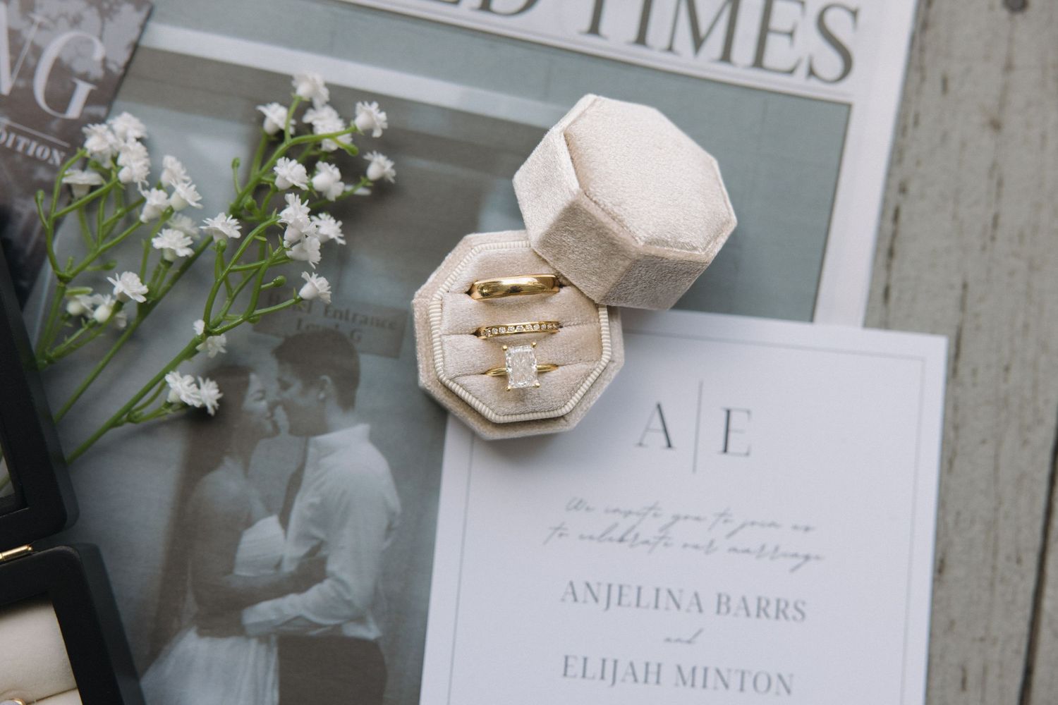 Wedding flat lay featuring gold rings in ivory velvet box, white invitation with monogram, and baby's breath flowers.