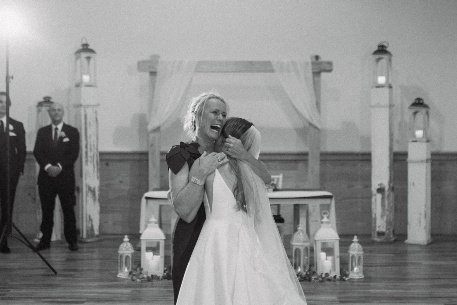 Black and white wedding photo of bride and groom sharing an emotional hug at the altar with lanterns and arch decor.